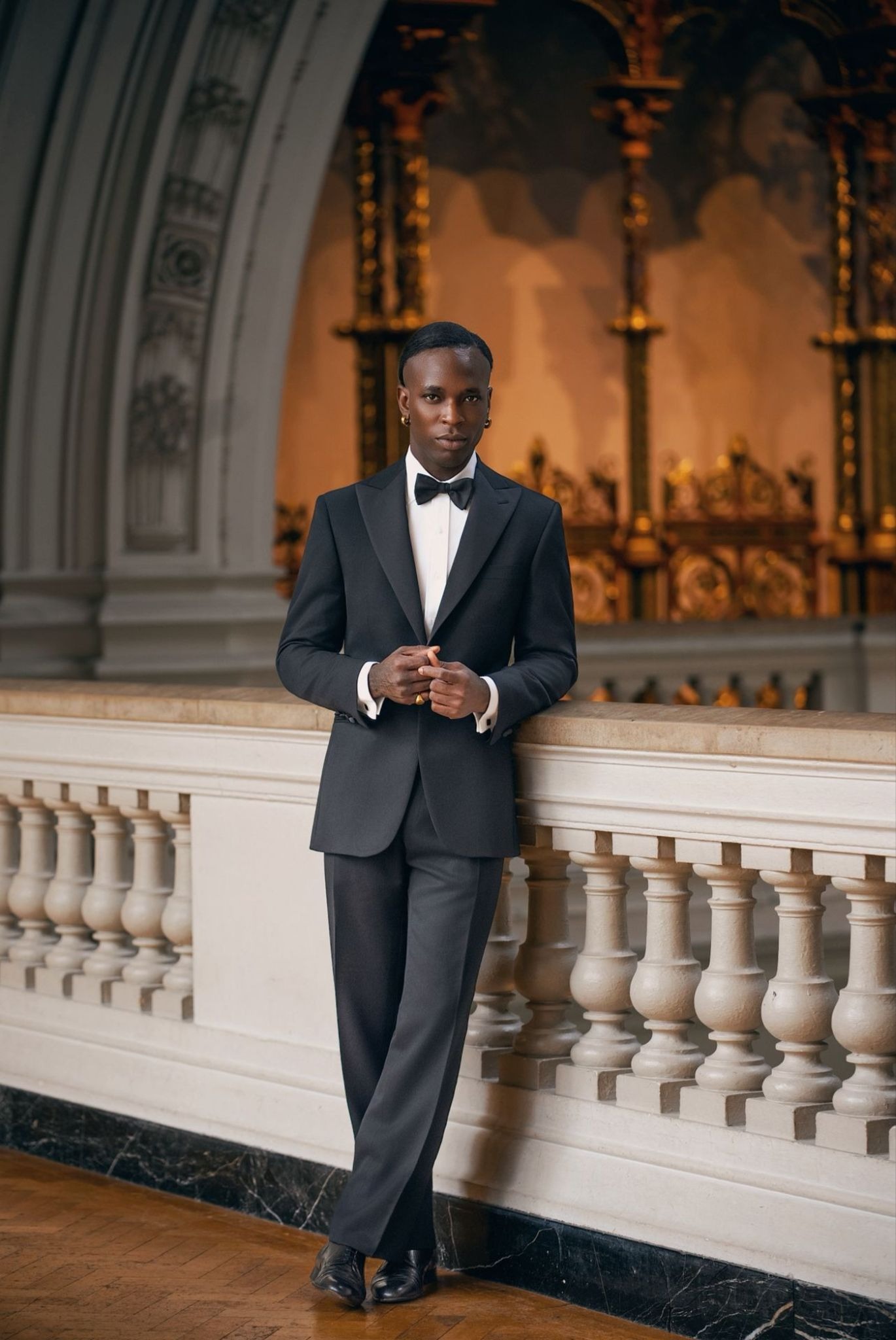 A person in a black tuxedo and bow tie stands elegantly against a white balustrade in an ornate, gold-accented hall, exuding sophistication and poise.