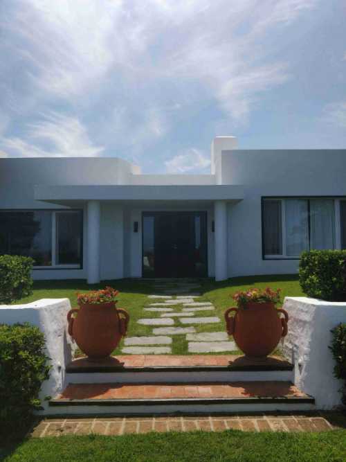 Stone path leads to a white modern house flanked by terracotta urns on brick steps under a wispy blue sky.