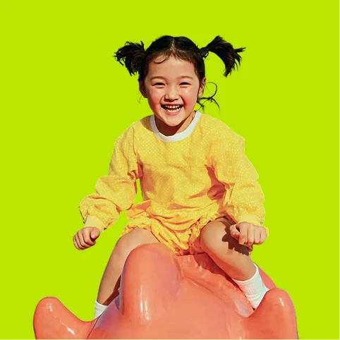 A young girl in a yellow outfit with pigtails smiles while sitting on a large pink sculpture, set against a bright green background.