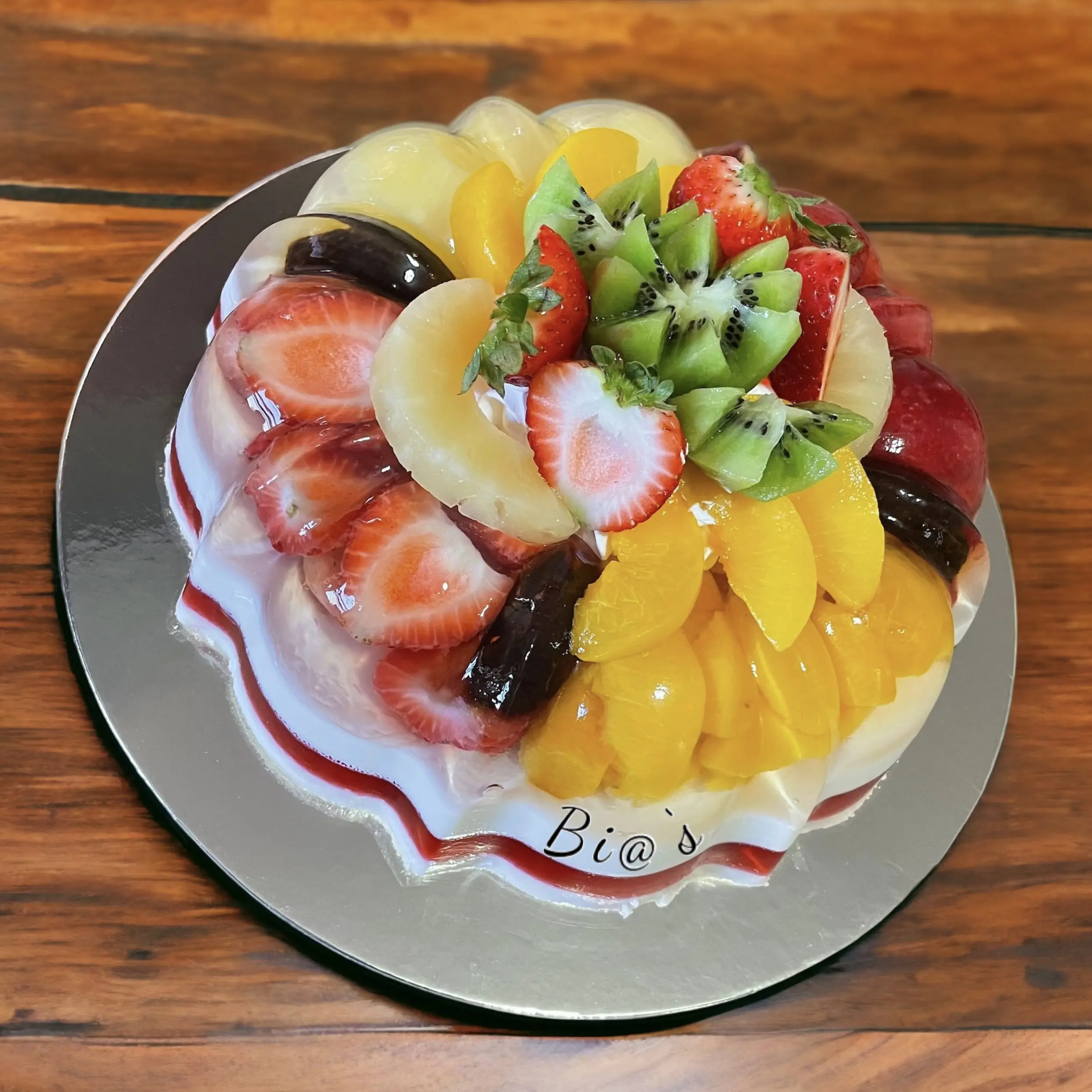 Fruit-topped gelatin cake featuring an array of fresh strawberries, kiwis, peaches, and pineapple slices arranged on a creamy base. A vibrant and refreshing dessert perfect for any gathering. Susa’s Sweets & Balloons.