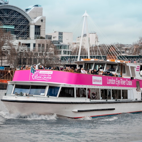 A crowded boat with a pink banner reading, "London Eye River Cruise," sails on a river with urban buildings and a bridge in the background.