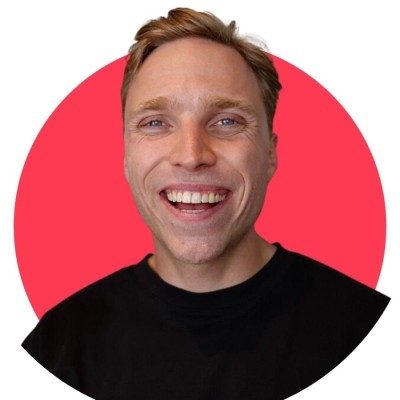 Smiling man in a black suit in a warmly-lit, modern cafe. Background features hanging lights and blurred people, creating a cozy, upscale ambiance.