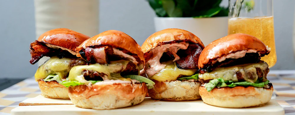 Four gourmet beef burgers on a wooden board, topped with melted cheese, crispy bacon, and lettuce in glossy brioche buns. A glass of sparkling drink sits in the background, creating a casual, indulgent meal setting.