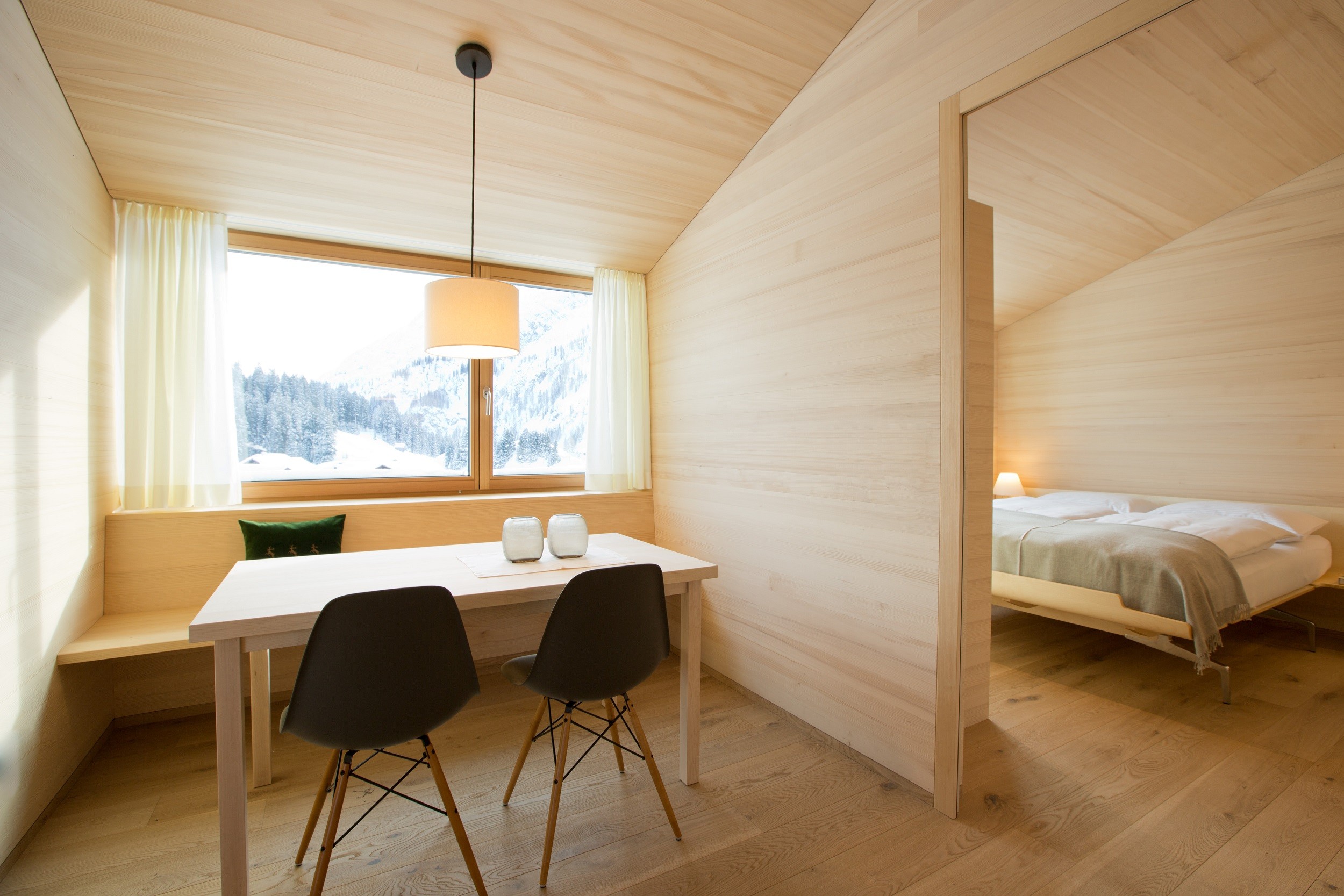 Minimalist alpine apartment with light-wood panelling, a small dining table with two chairs, and a bedroom visible through an open doorway, overlooking a snowy mountain landscape.