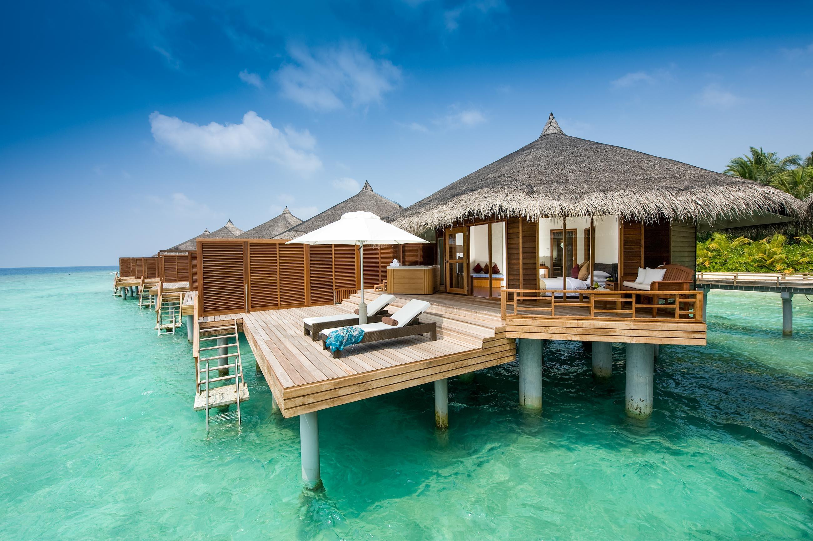 Overwater wooden bungalow with thatched roof and a sunny deck over turquoise ocean water in the Maldives.
