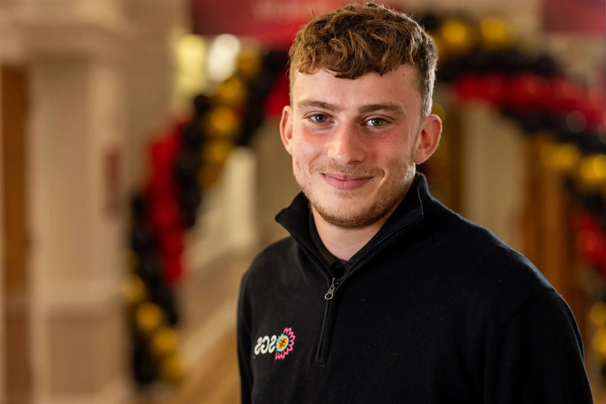 A young man with short hair smiles, wearing a black shirt, in a warmly decorated indoor setting.