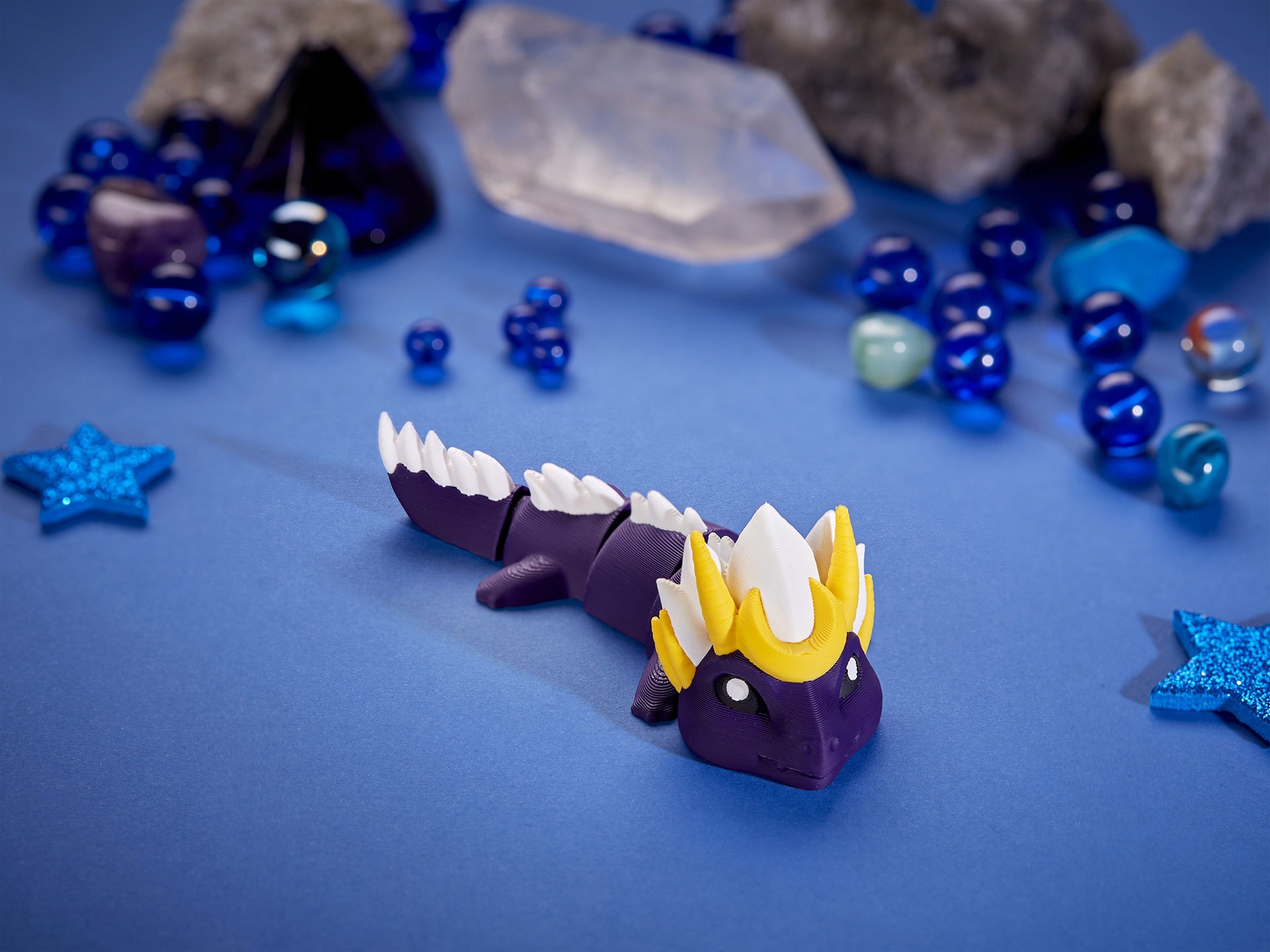 A 3D printed purple dragon figurine with a yellow crown and white spikes is surrounded by blue marbles and star decorations on a textured blue background.