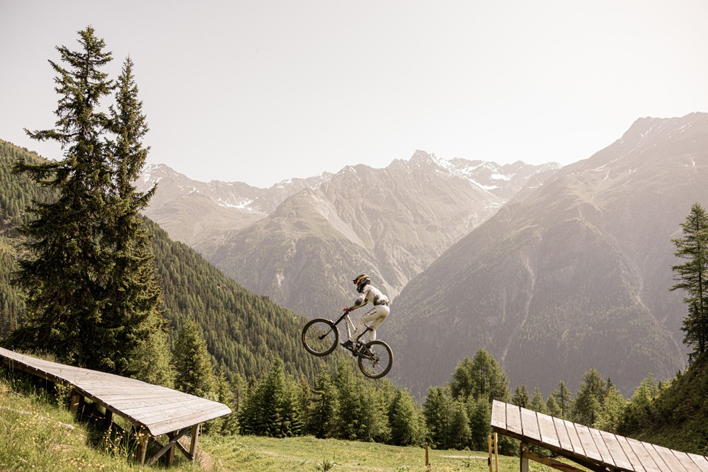 Mountainbiker in weißer Ausrüstung springt in einer immergrünen Baumlandschaft von einer Rampe.
