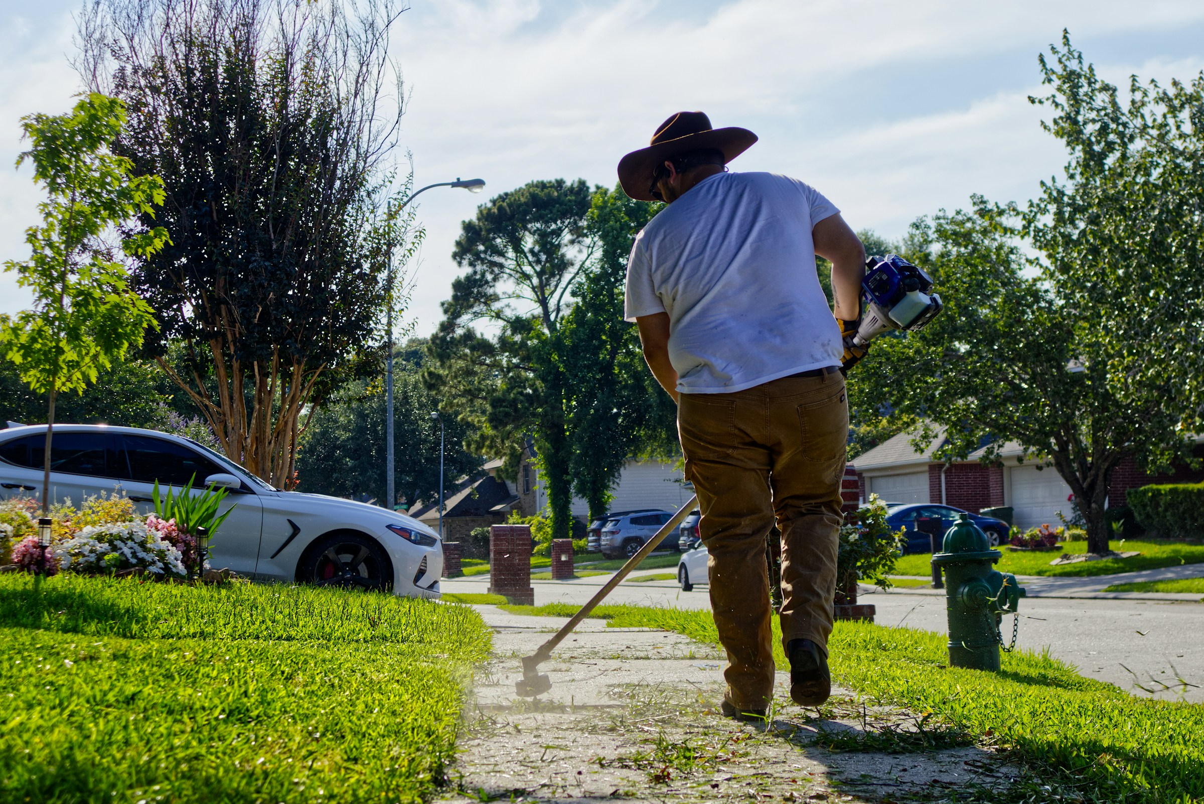 A landscaper using a string trimmer to edge a concrete sidewalk next to a manicured residential lawn.