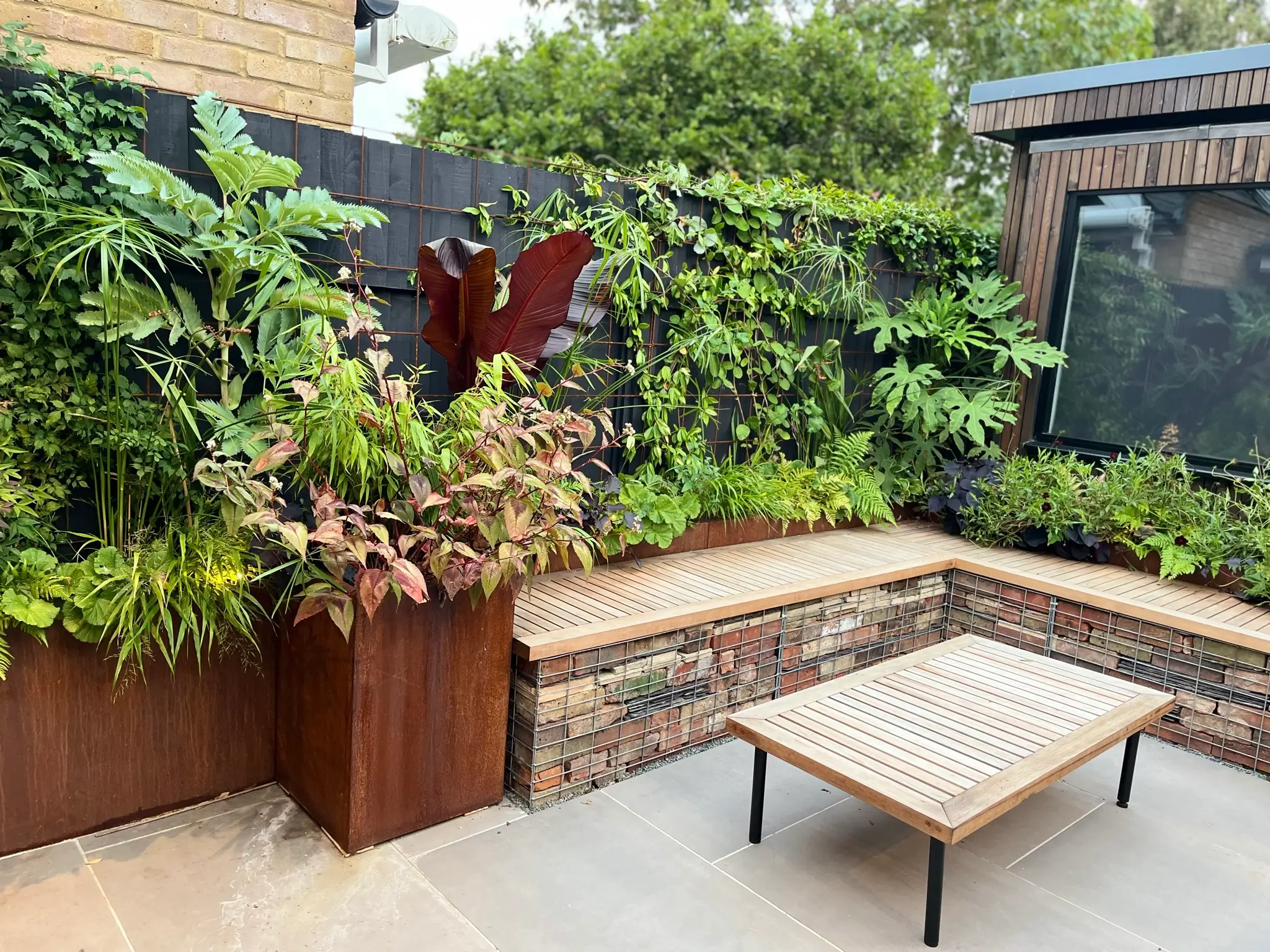 A cozy outdoor space with lush greenery, stone seating, and a wooden table set against a garden backdrop.