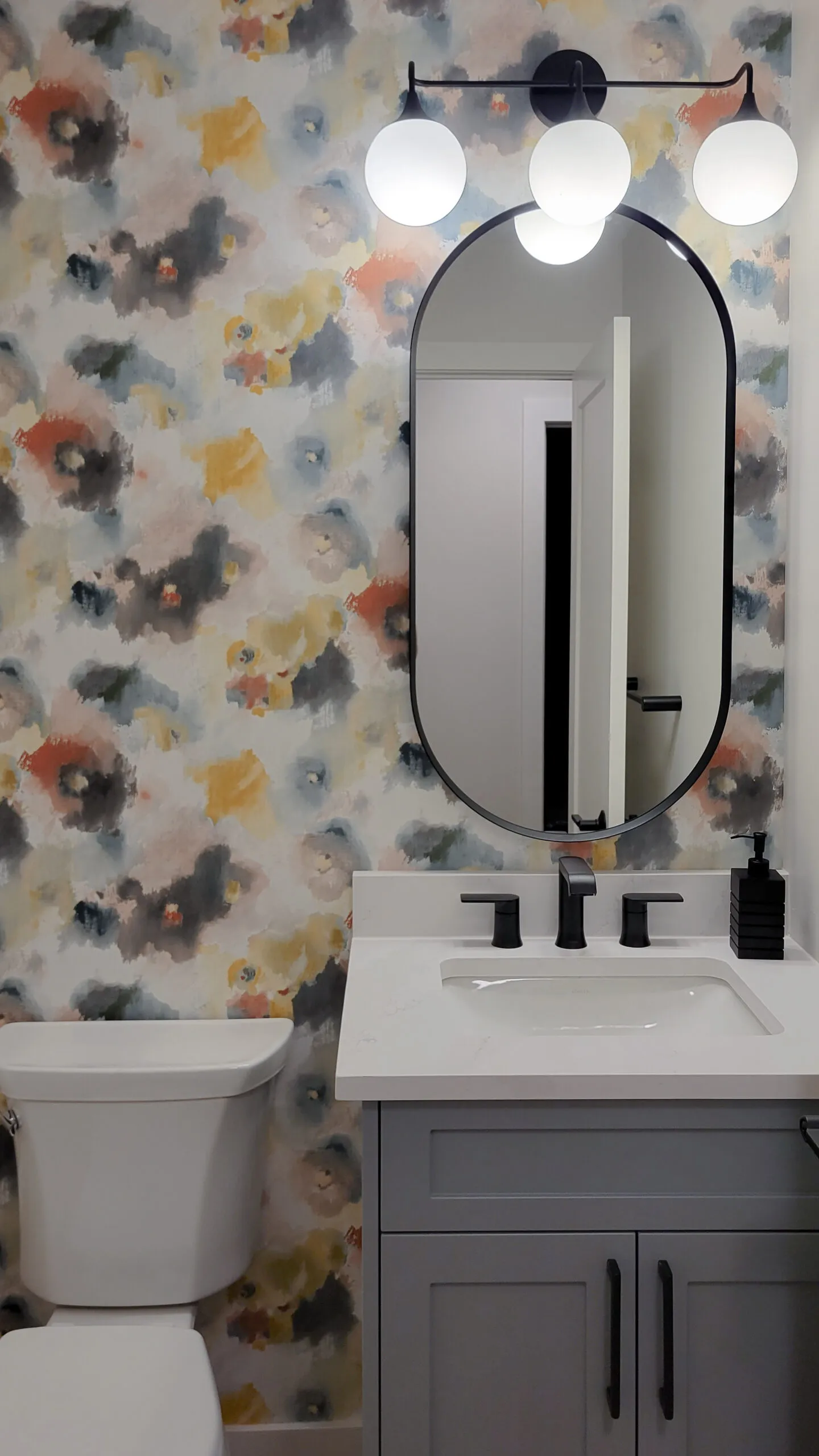 Image of powder room vanity with colour splotch wallpaper and black racetrack shaped mirror.