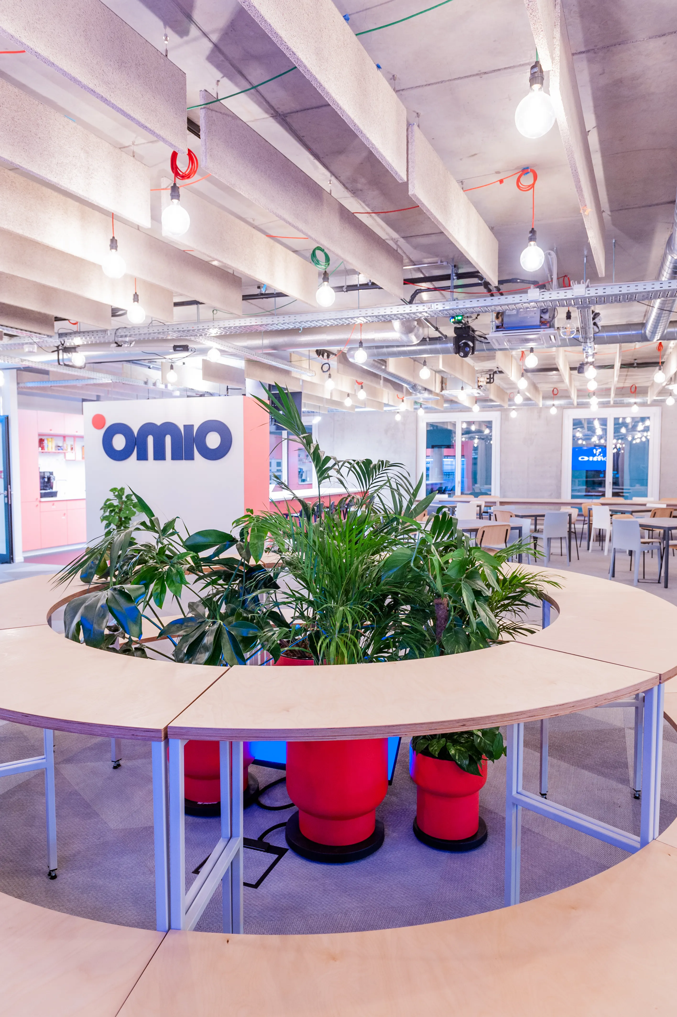Circular plywood standing table at OMIO with a planted island of palms and ferns in red and blue pots at its centre, beneath an exposed concrete ceiling with bare bulbs on colourful hooks.