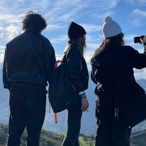 Three people in winter clothing stand on a hill, one taking a photo with a smartphone, with mountains and clouds in the background.