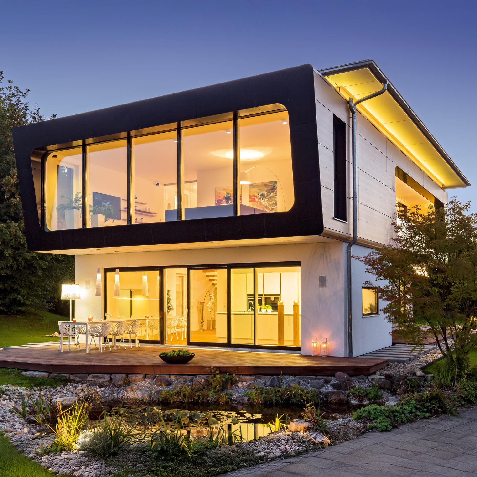 Modernes zweistöckiges Haus mit großflächigen Glasfronten und einer Terrasse am Abend. Im Vordergrund ein kleiner Teich mit Beleuchtung.