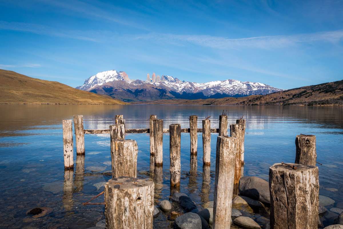 Wooden pier posts stand in clear blue water in front of snow-capped mountains under a bright blue sky.