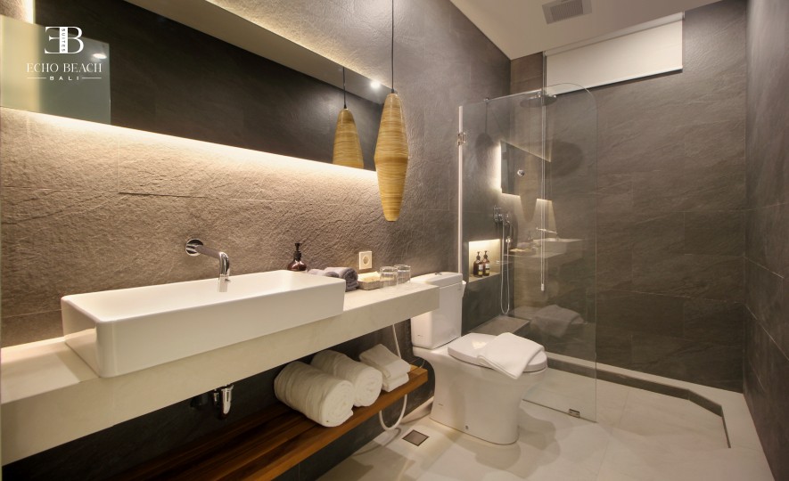 Bali Two Bedroom Bathroom: Modern ensuite with dark stone tiles, illuminated mirror, white sink, and wood storage shelf. Luxury walk-in shower at Echo Beach Suite, Canggu.