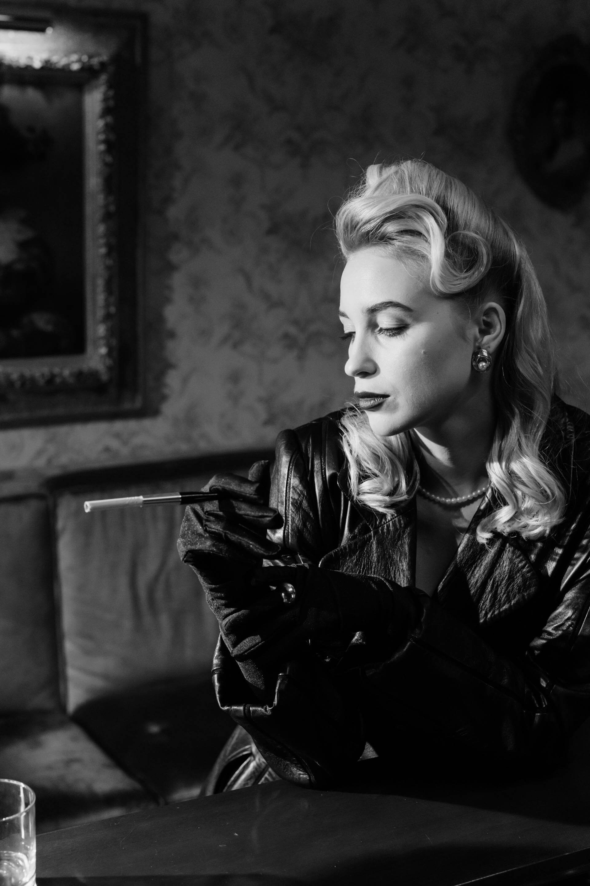 A woman in a leather jacket sits at a table, holding a cigarette, exuding a confident and relaxed demeanor