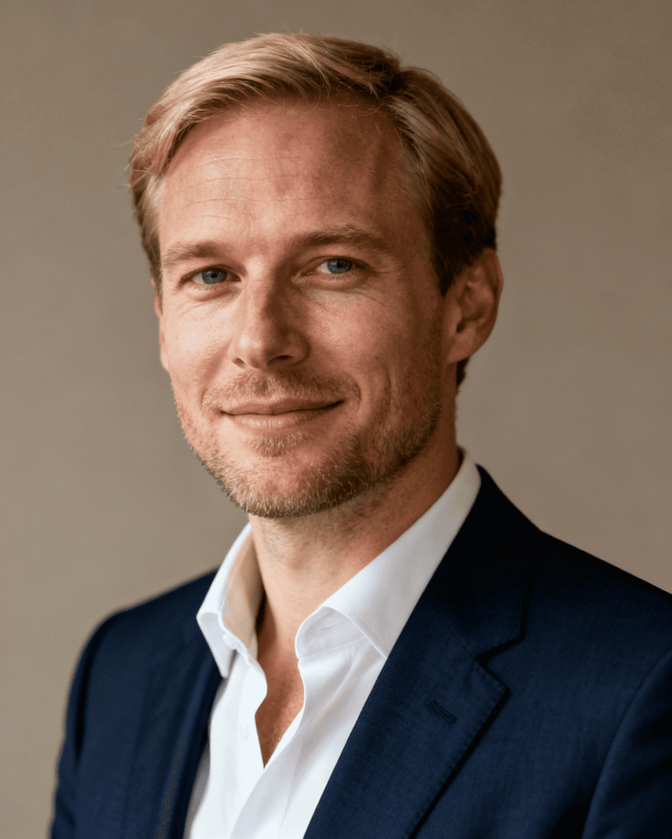 A man with blonde hair and light stubble smiles gently in a professional headshot. He's wearing a navy suit and white shirt against a neutral background.