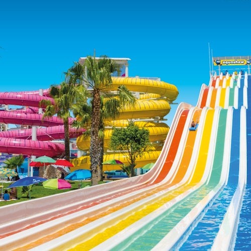 Toboganes de agua coloridos, palmeras y sombrillas bajo un cielo azul brillante en un parque acuático.