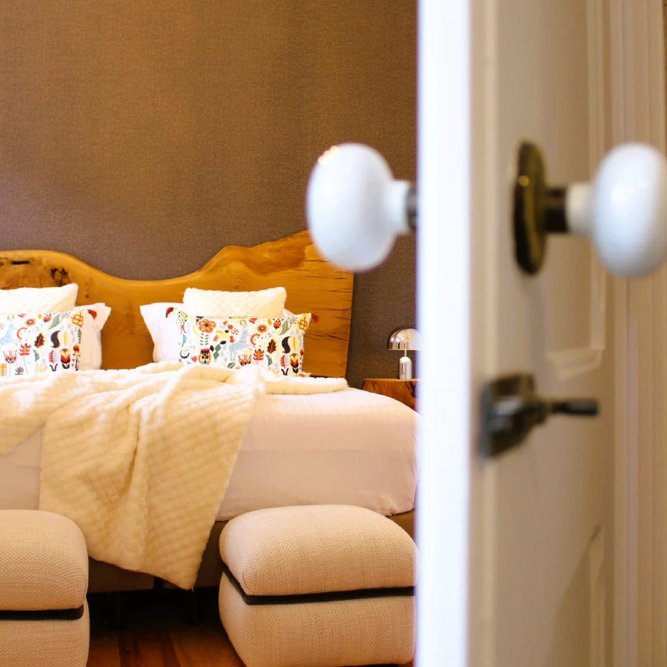 Cozy bedroom with a wooden headboard and colorful floral pillows on a neatly made bed, seen through an open door. Soft lighting creates a warm ambiance.