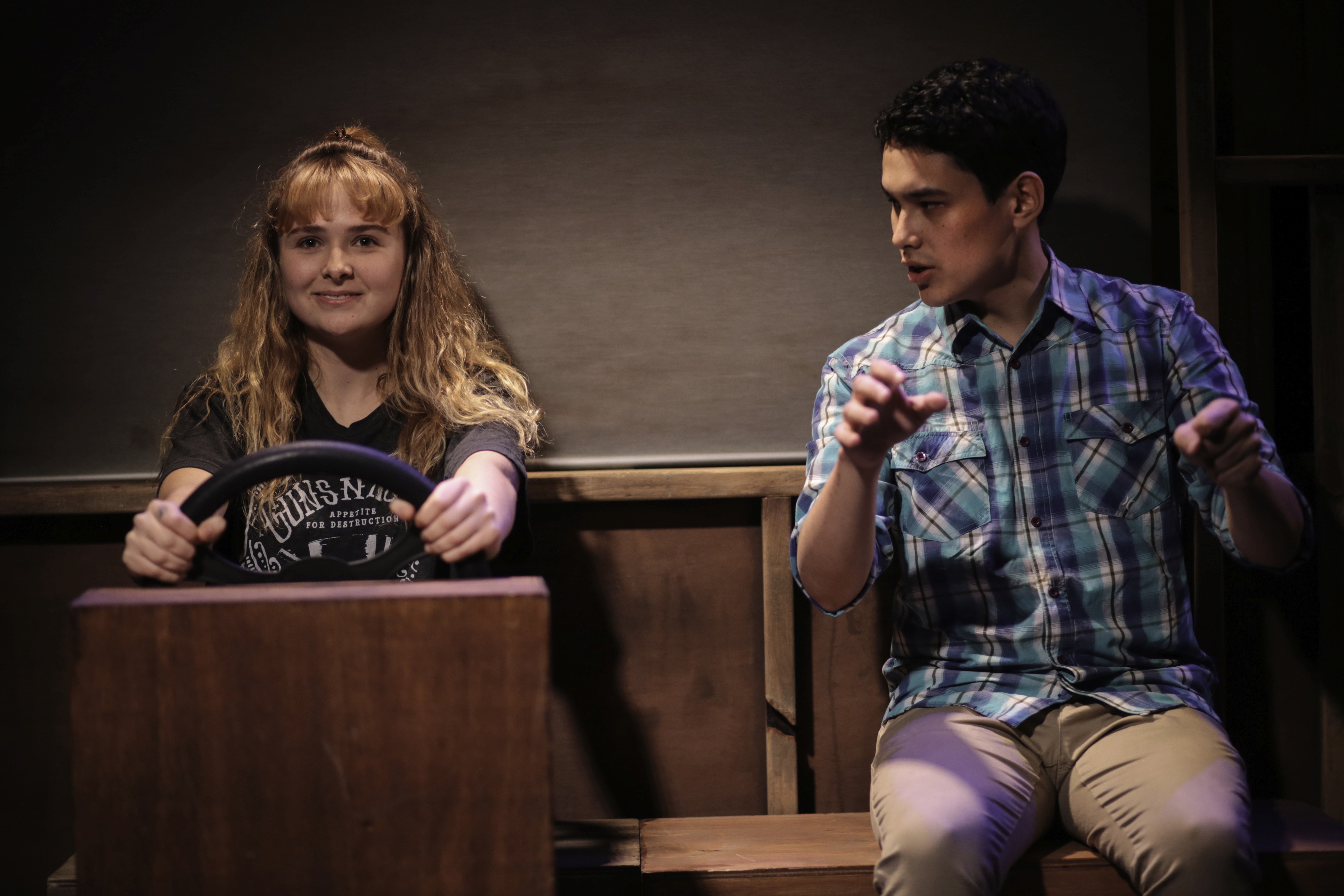 A girl sits behind a wooden steering wheel prop, smiling, while a young man beside her mimes driving gestures. Their playful expressions contrast with the minimal stage design.
