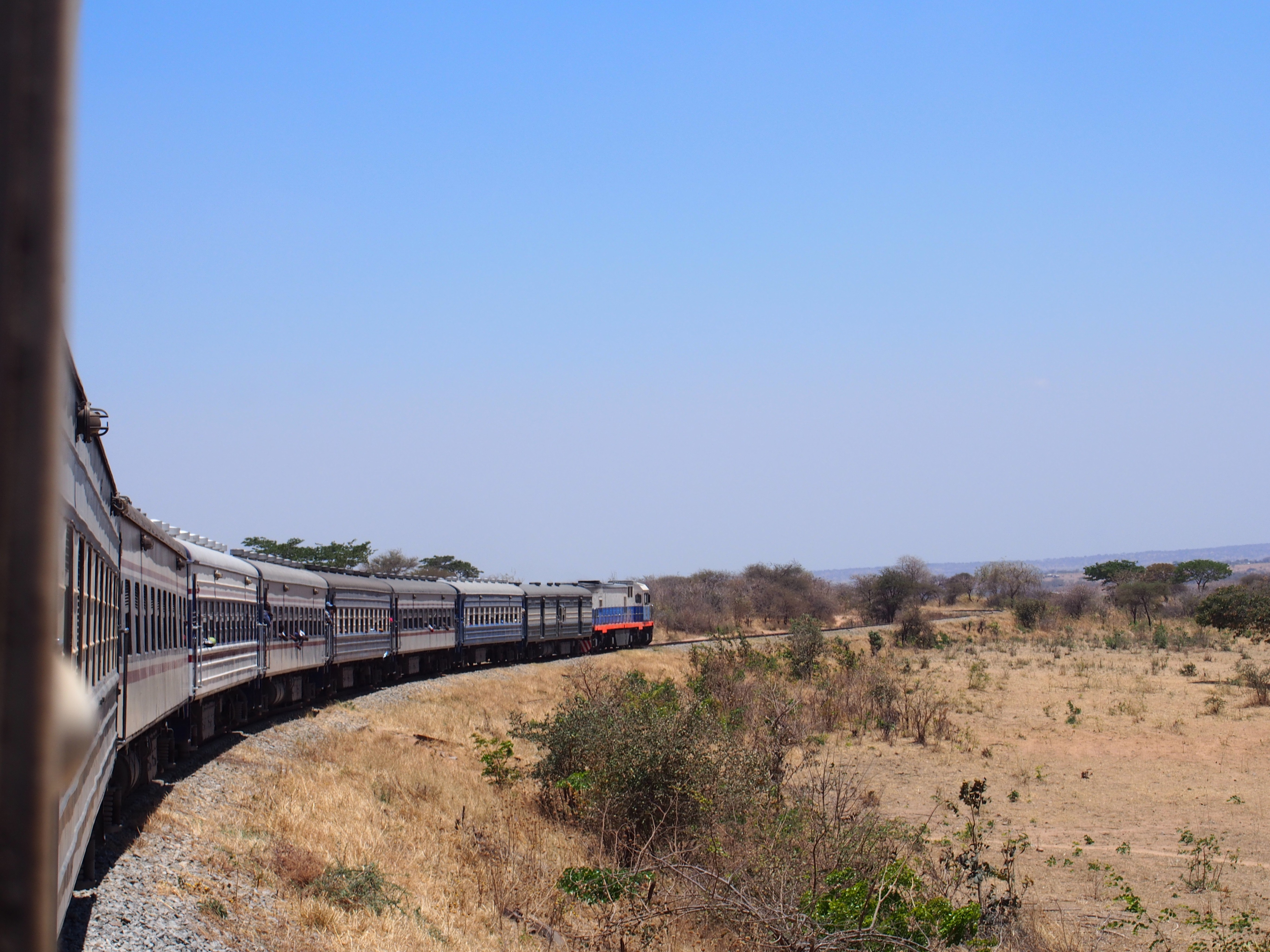 A long passenger train curves through a dry, open savanna landscape under a clear blue sky.