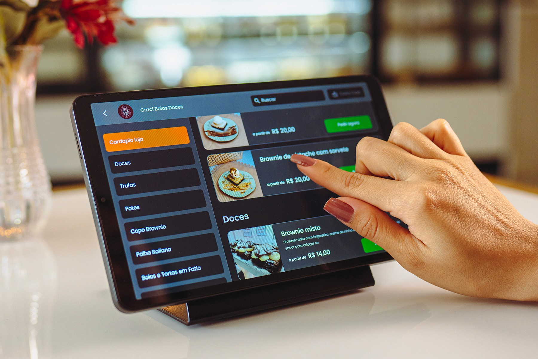 Pessoa utilizando tablet para visualizar menu de doces, com destaque para opções de bolos e brownies em uma confeitaria.
