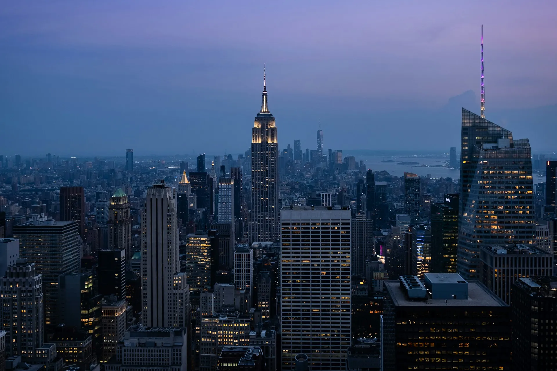New York City skyline at twilight featuring the Empire State Building and other skyscrapers illuminated against a purple-blue sky.