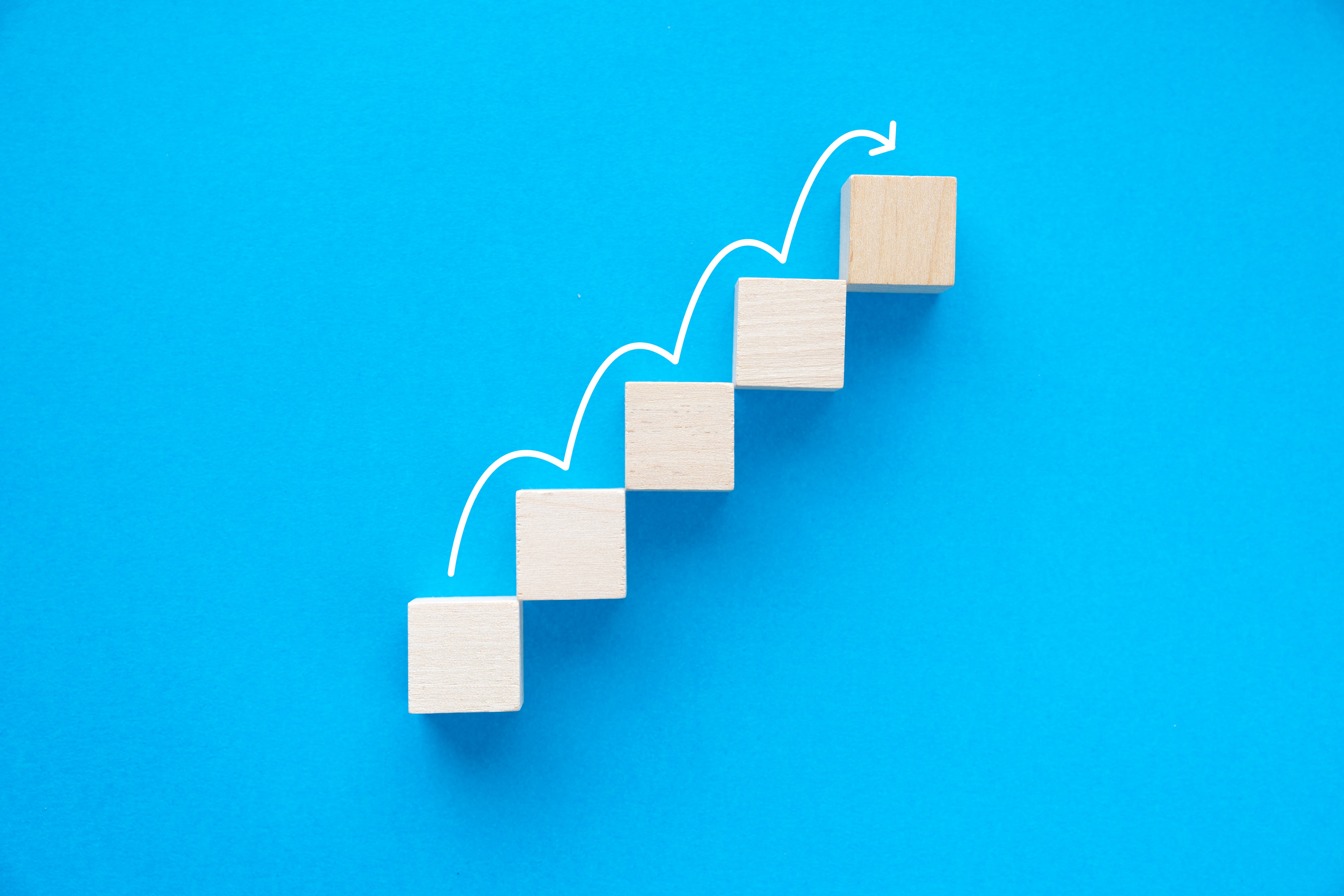 Business growth concept with wooden blocks forming upward steps and white arrow on blue background.