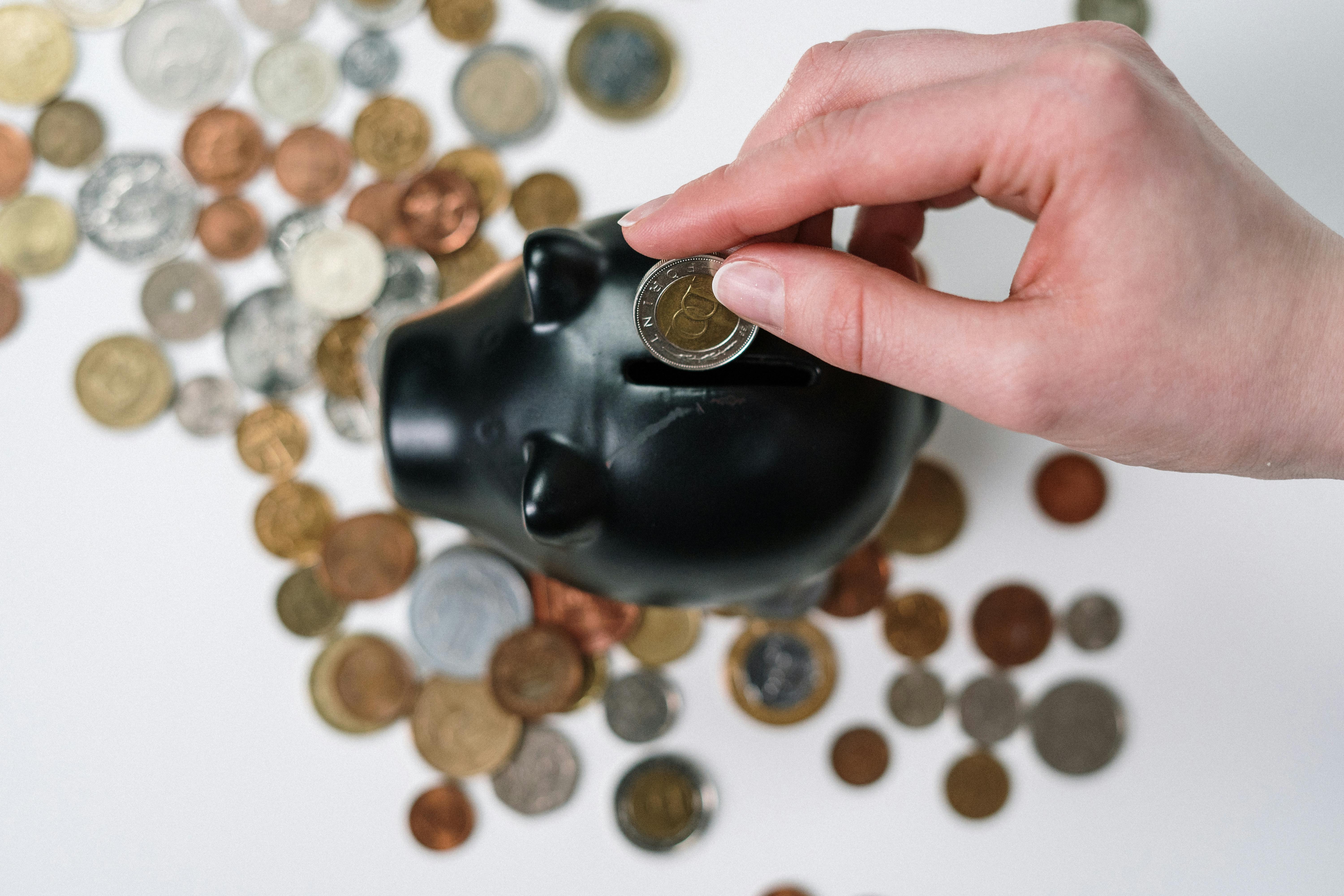 A hand placing a coin into a black piggy bank surrounded by various coins on a white surface.