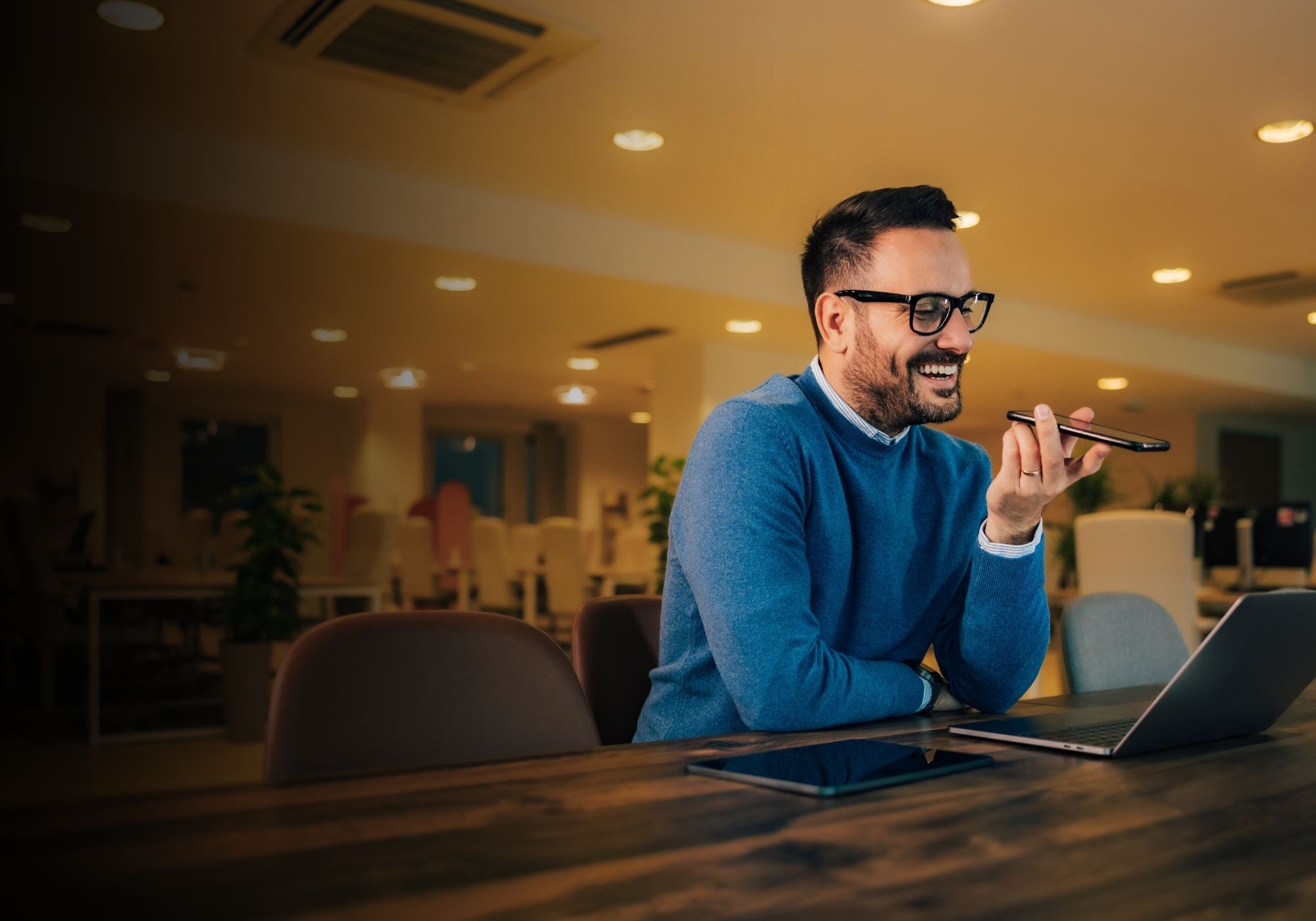Man in glasses wearing a green shirt, writing in a notebook at a speckled table with a cup of coffee nearby, engaged in conversation.