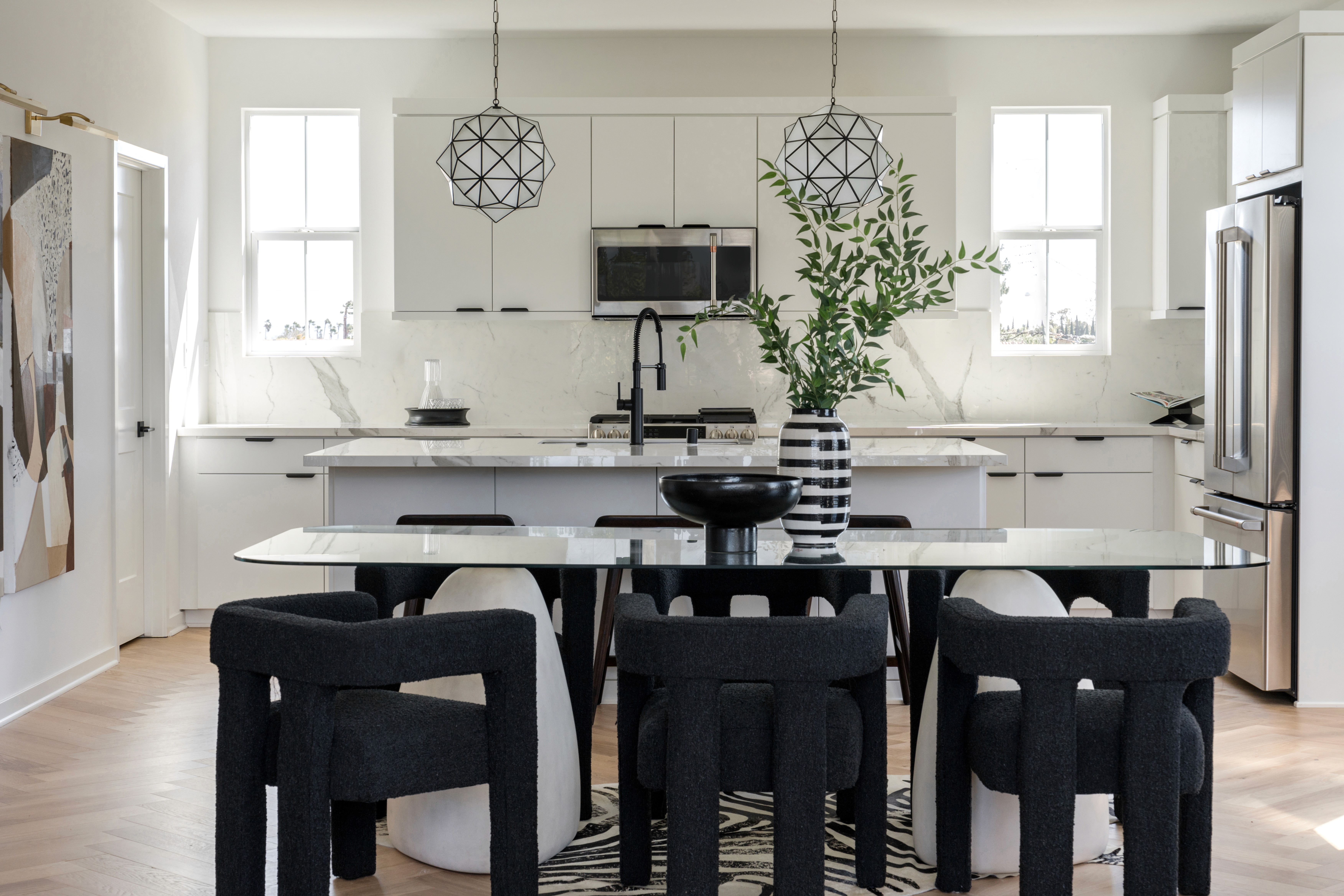 Dining area with black boucle dining room chairs, modern, glass top dining table, black pedestal bowl and black and white striped vase; kitchen in background with flat white cabinets, black hardware, white marbled countertops