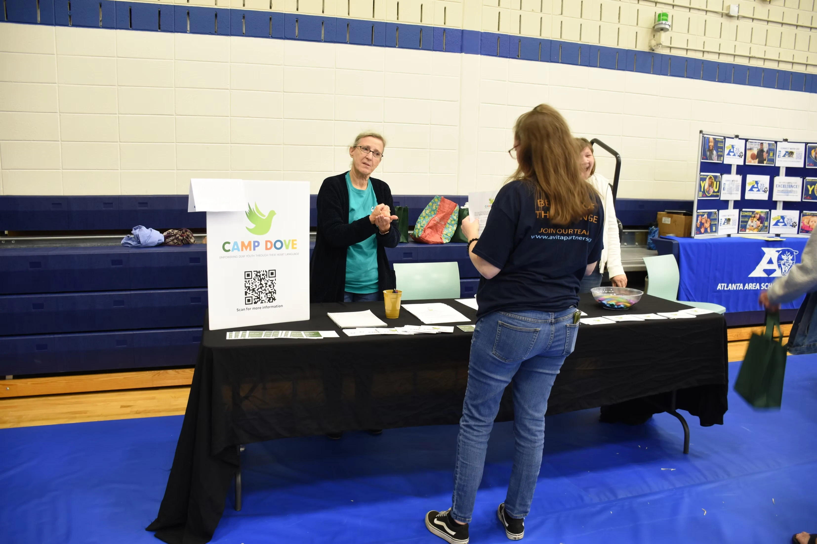 A woman at a table with informational pamphlets talks to a woman at a school event. A QR code is visible on the table.