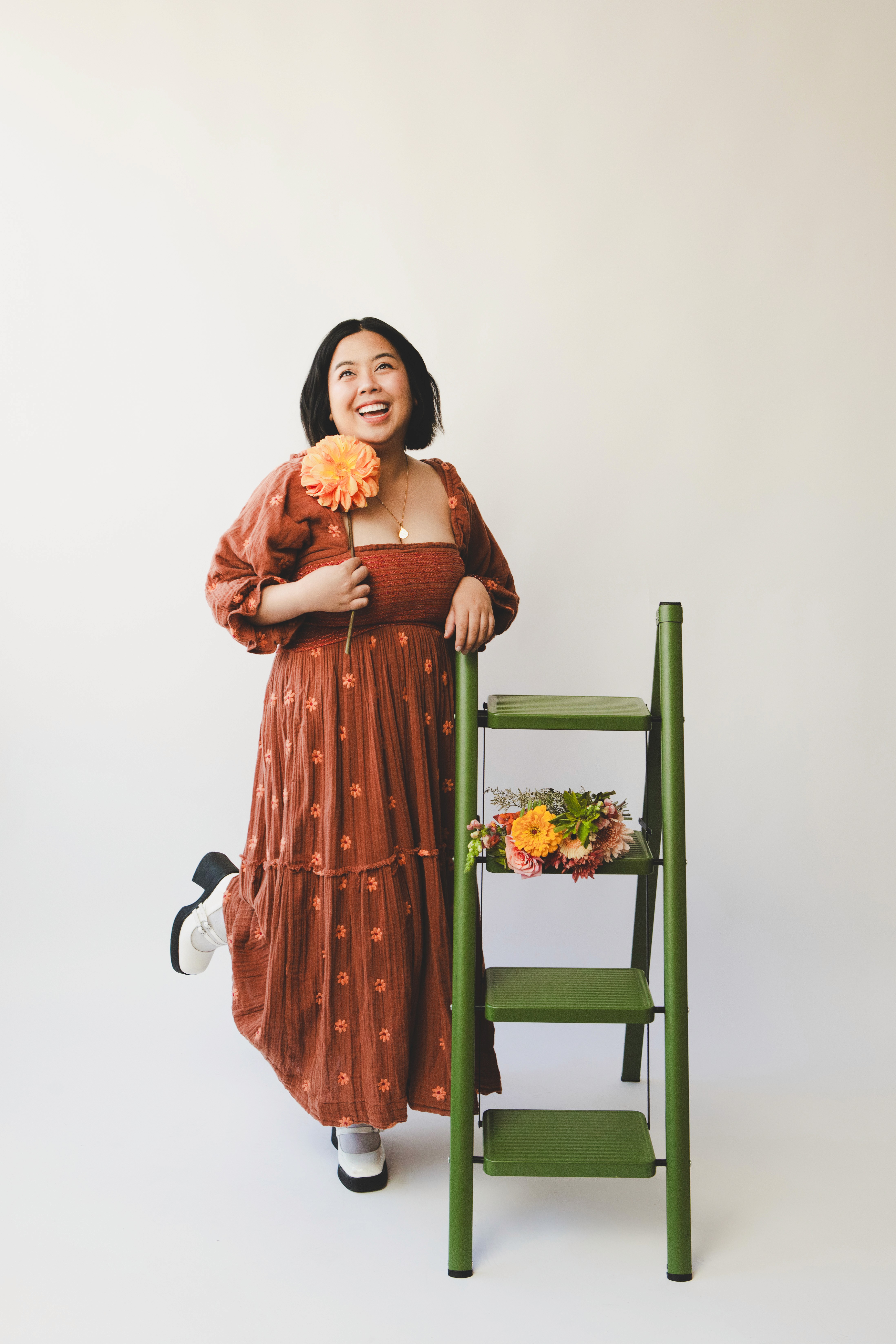 designer holding single yellow flower as she leans against green ladder