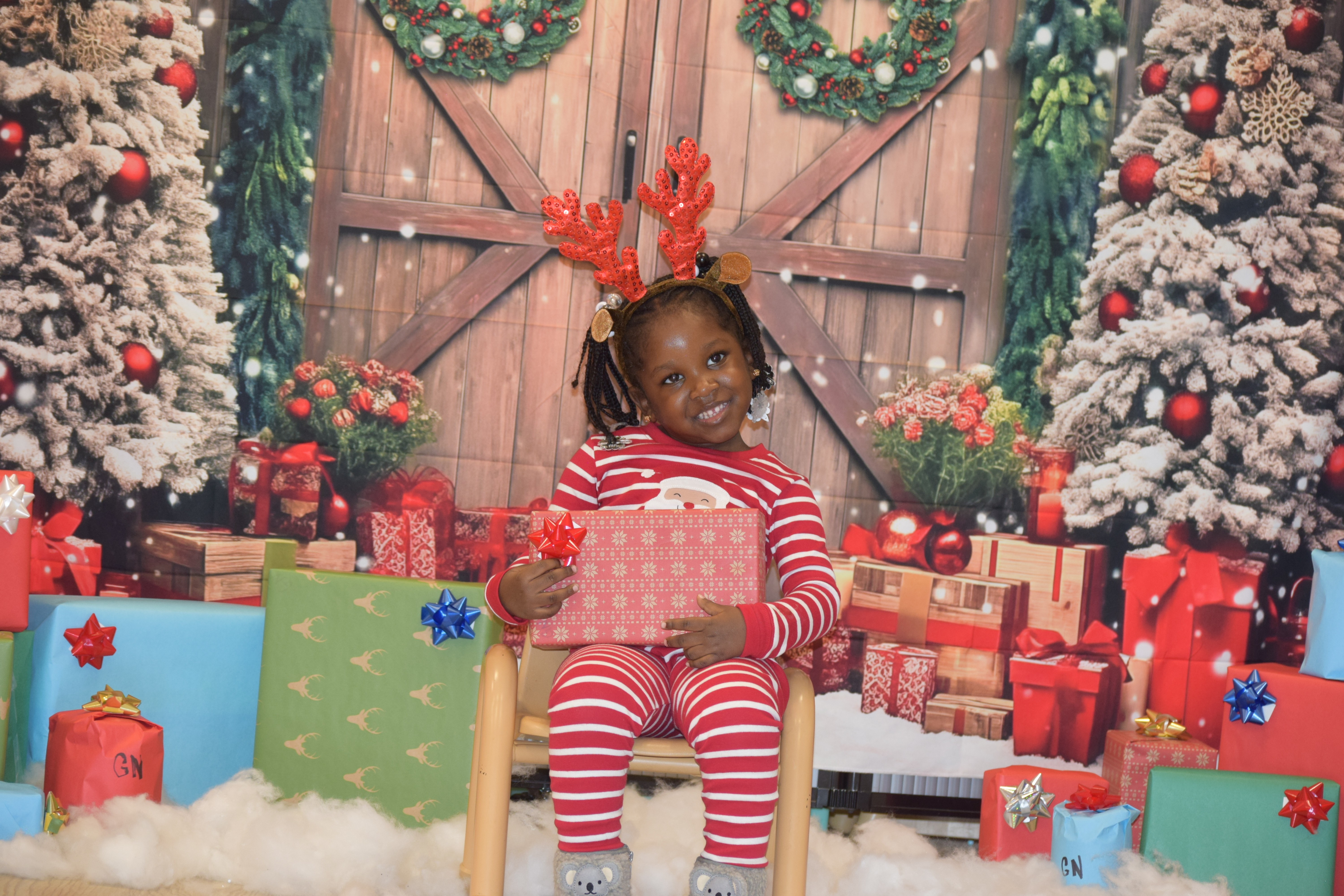 A young kid in Christmas pajamas and reindeer antlers holds a present in a Santa's Workshop inspired background. 