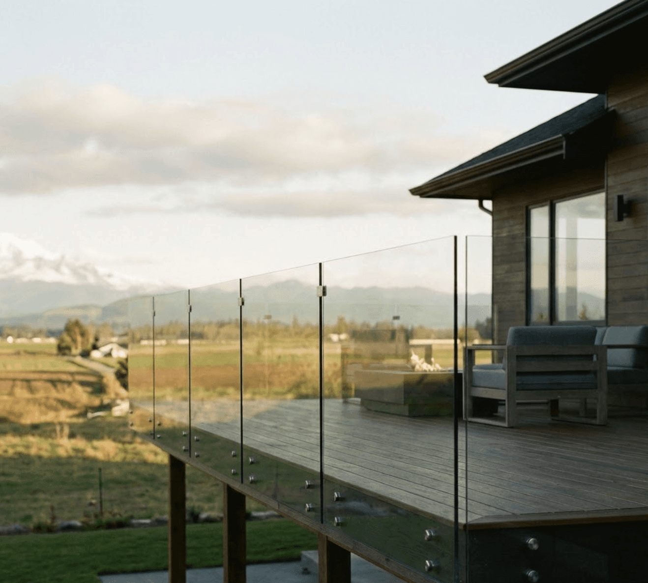 A stunning modern backyard deck with frameless glass railings in Abbotsford, British Columbia. View overlooks farmland and Mount Baker in the distance. Golden hour lighting with soft Pacific Northwest clouds. The deck is attached to a contemporary Fraser Valley home with dark wood decking. Clean, unobstructed glass panels with minimal hardware.