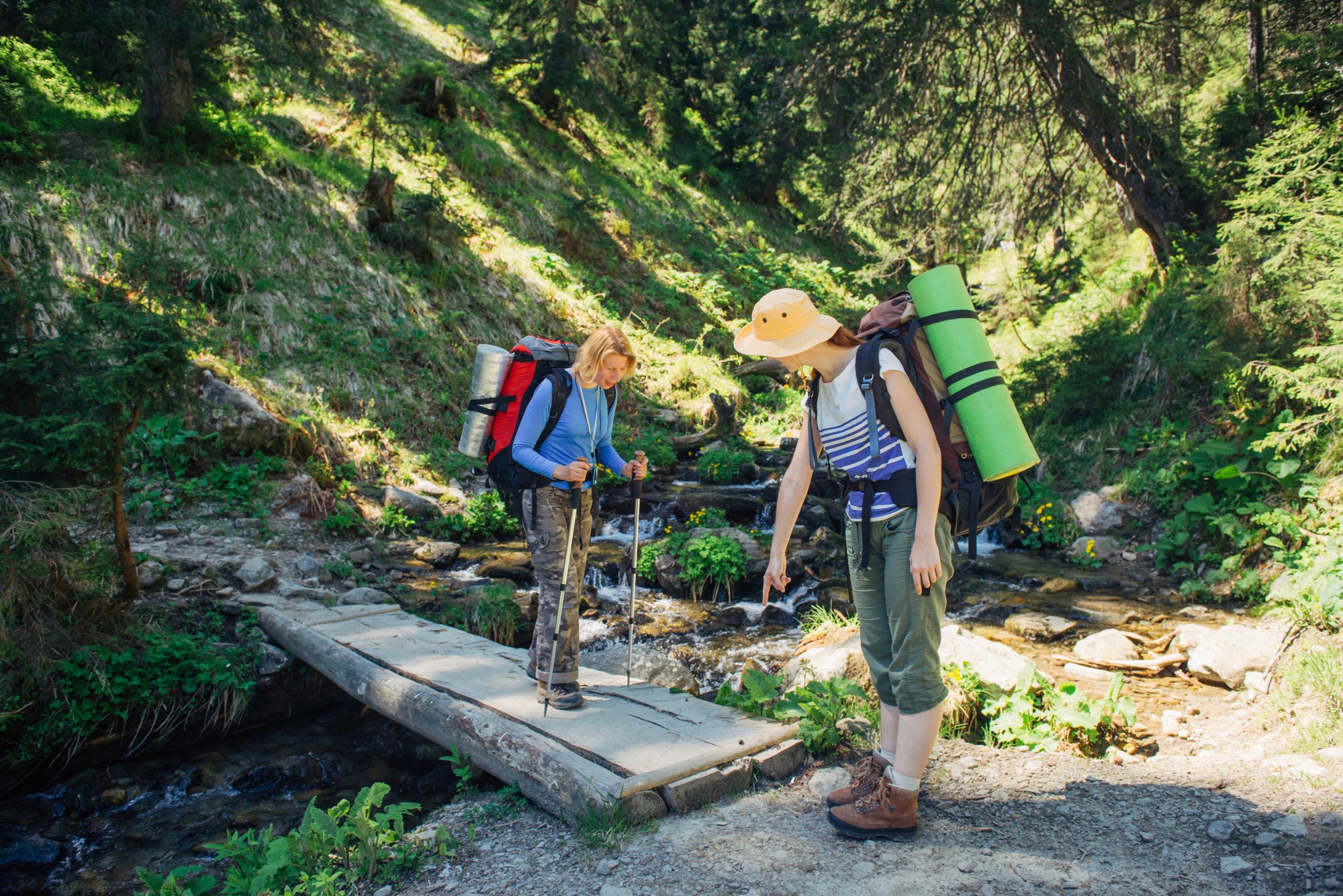Two female hikers with large backpacks and trekking poles standing on and near a rustic wooden bridge over a small mountain stream in a sunlit forest.