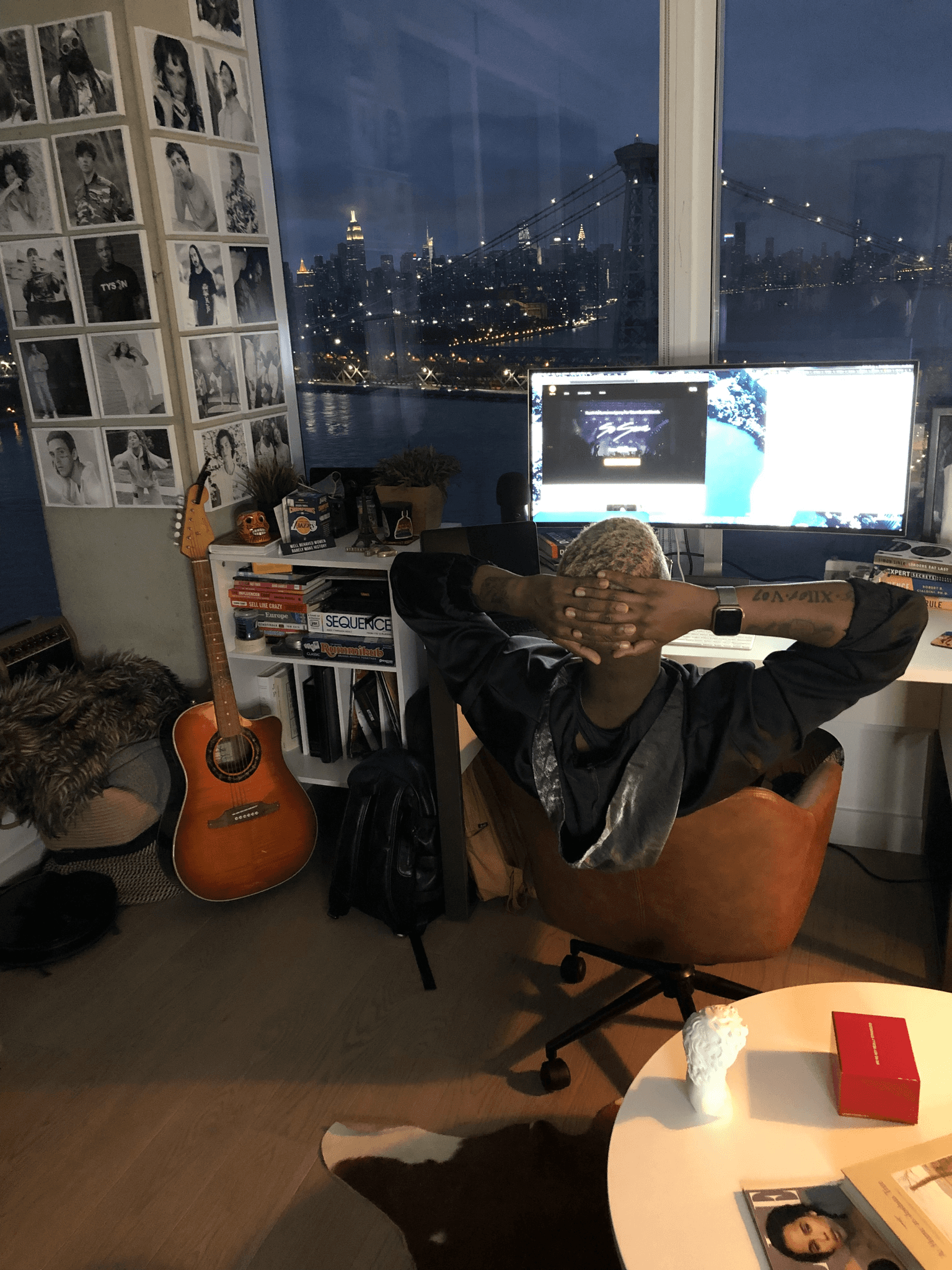 A cozy workspace with a computer, guitar, and a view of a city skyline at dusk.