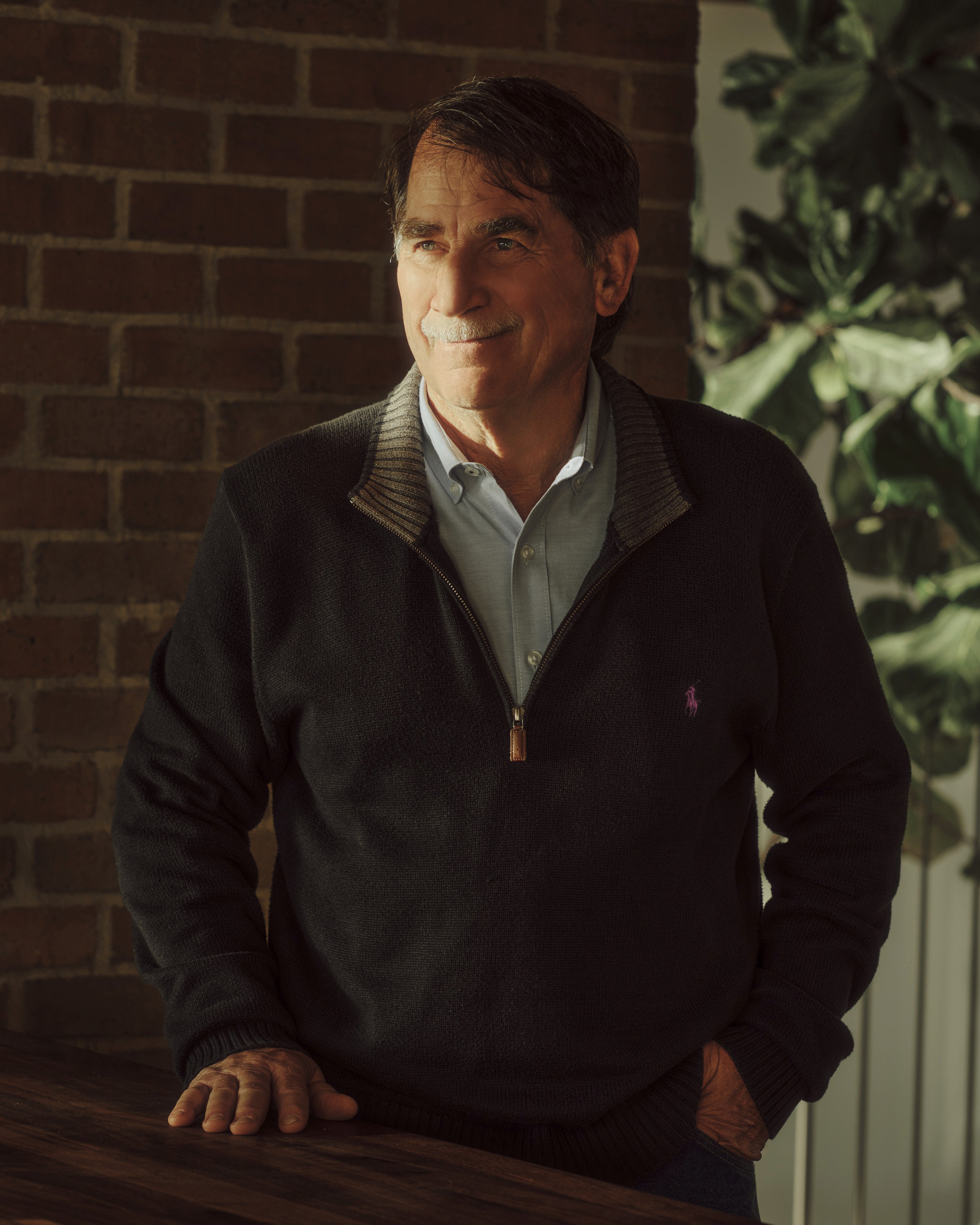 A professional portrait of a smiling man with a mustache, wearing a quarter-zip sweater, leaning against a wooden table in front of a brick wall.