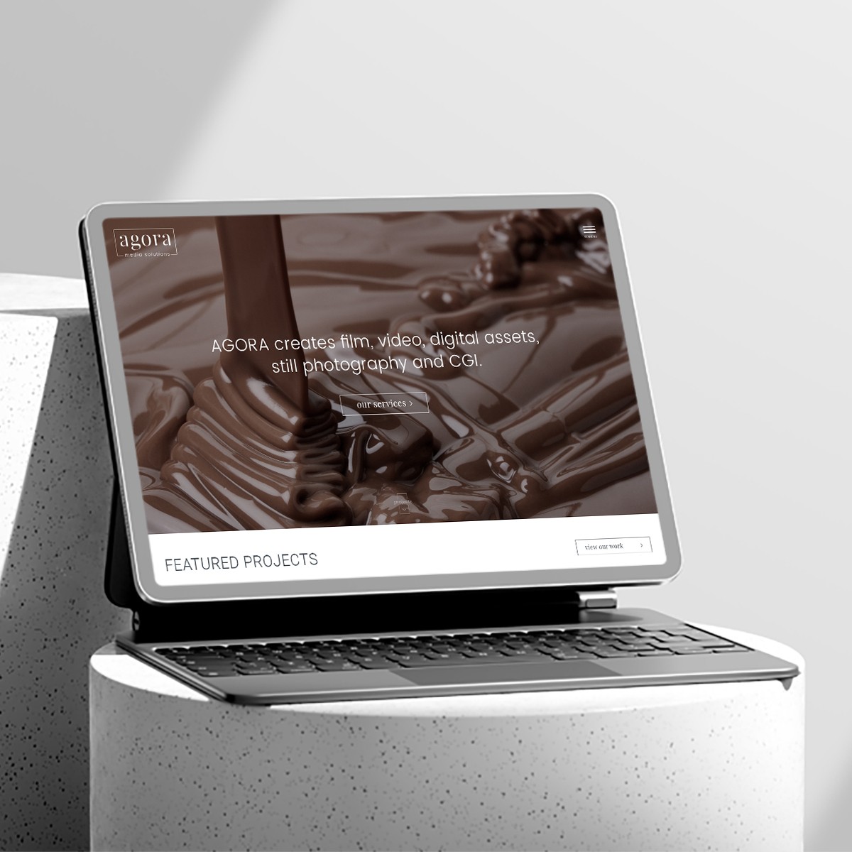 Thumbnail image of a laptop on concrete stand displaying Agora Media Solutions homepage with chocolate visual and featured projects section.