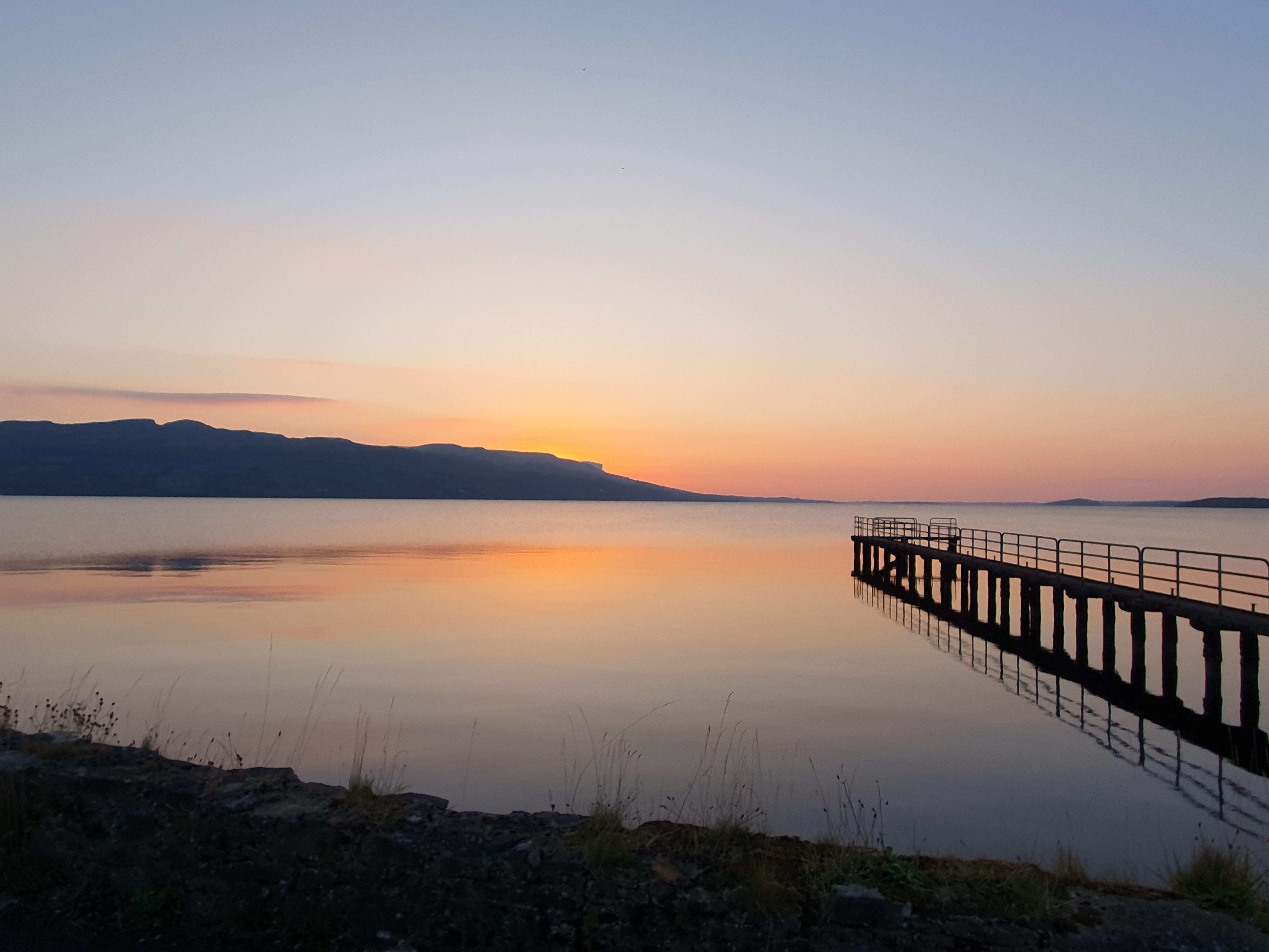 Serene sunset over calm waters, with a wooden pier extending into the lake and mountains in the background.