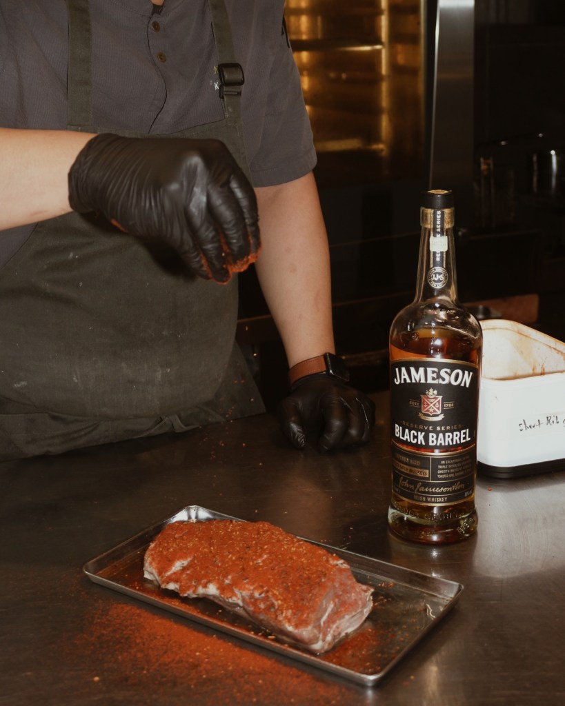 A chef in black gloves sprinkling seasoning onto a piece of meat on a metal tray, with a bottle of Jameson Black Barrel whiskey nearby.