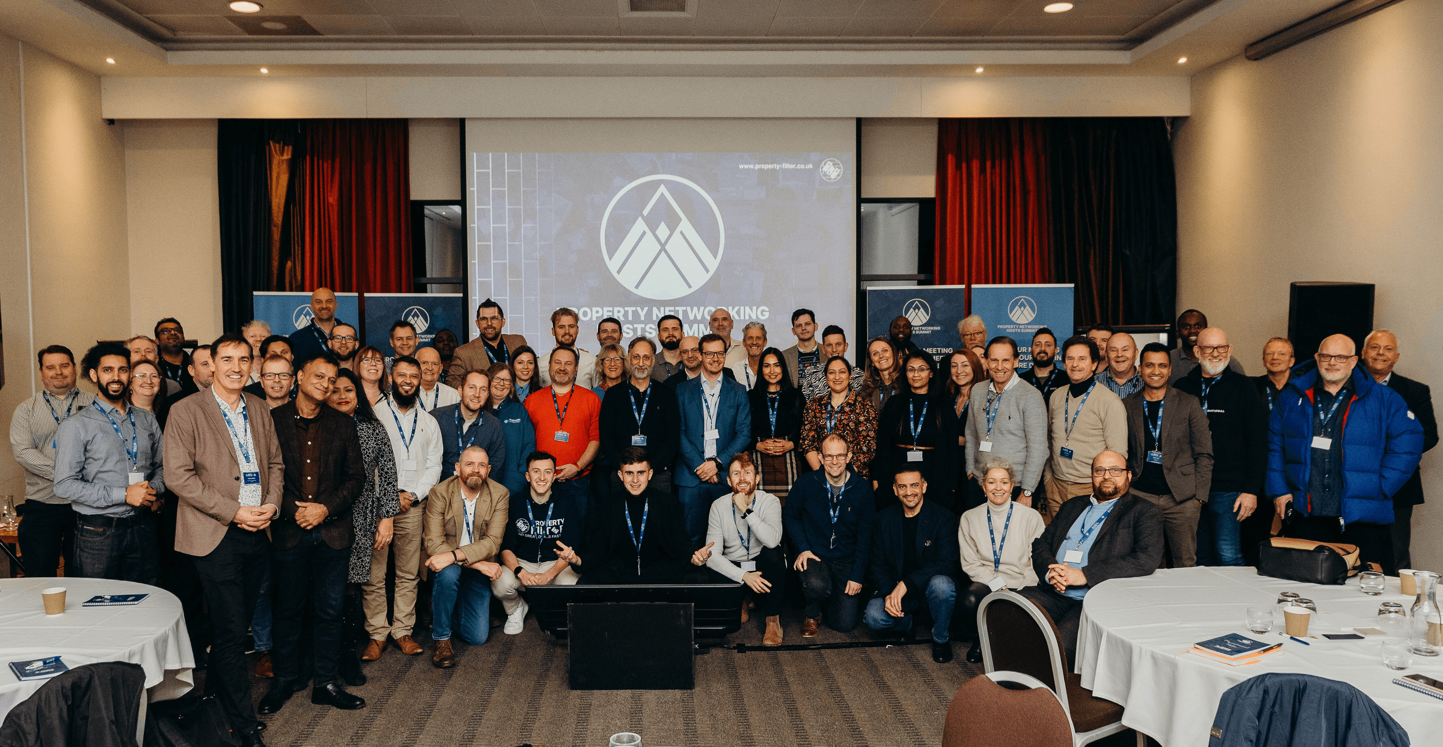 Large group photo of approximately 60-70 attendees at Property Networking Hosts Summit conference, standing in tiered rows in conference room with projection screen showing mountain logo