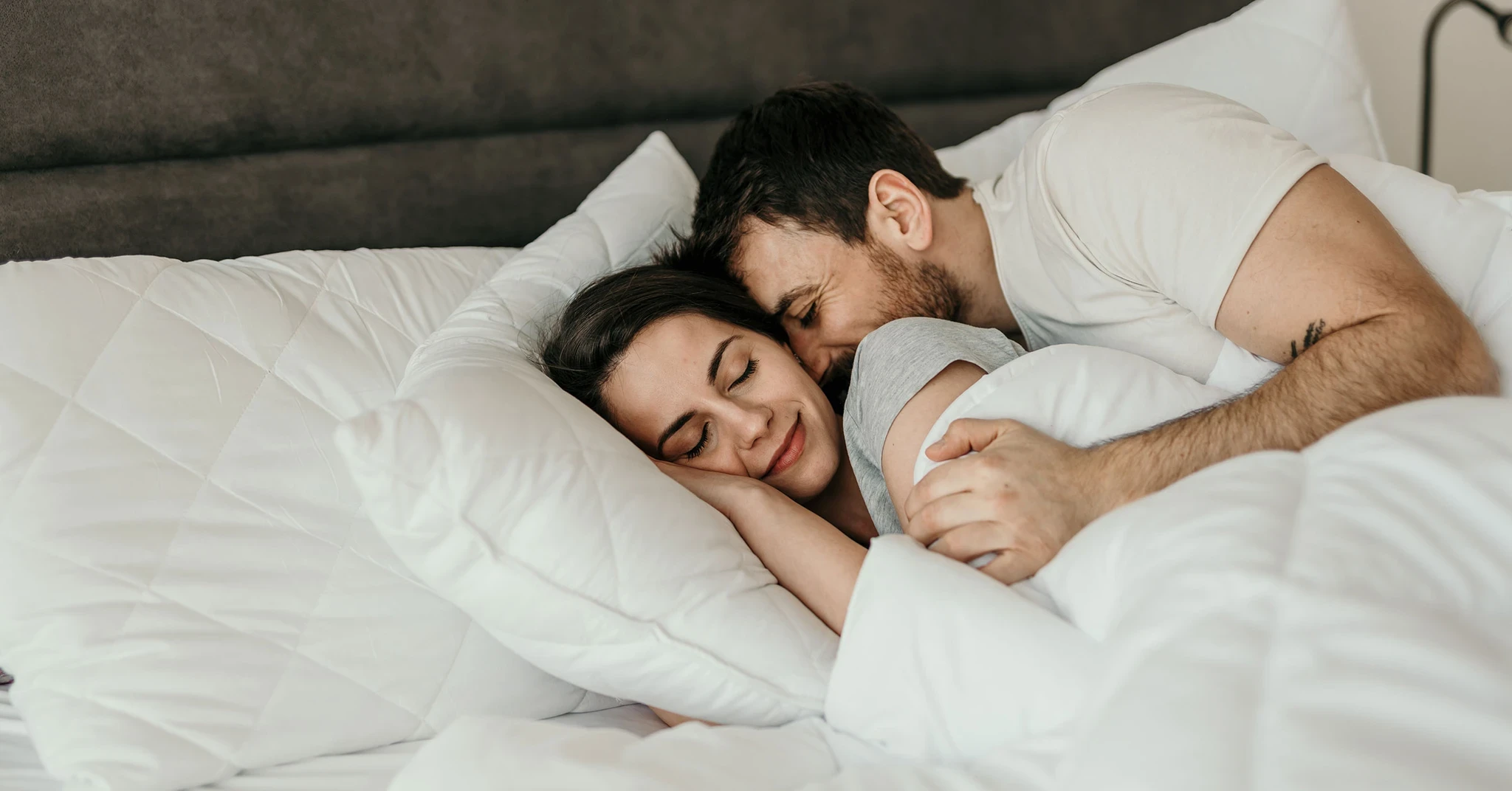 Ett par ligger tätt intill varandra i en säng. Mannen omfamnar kvinnan bakifrån. De verkar befinna sig i en lugn och kärleksfull stund, med slutna ögon och ett leende på läpparna.