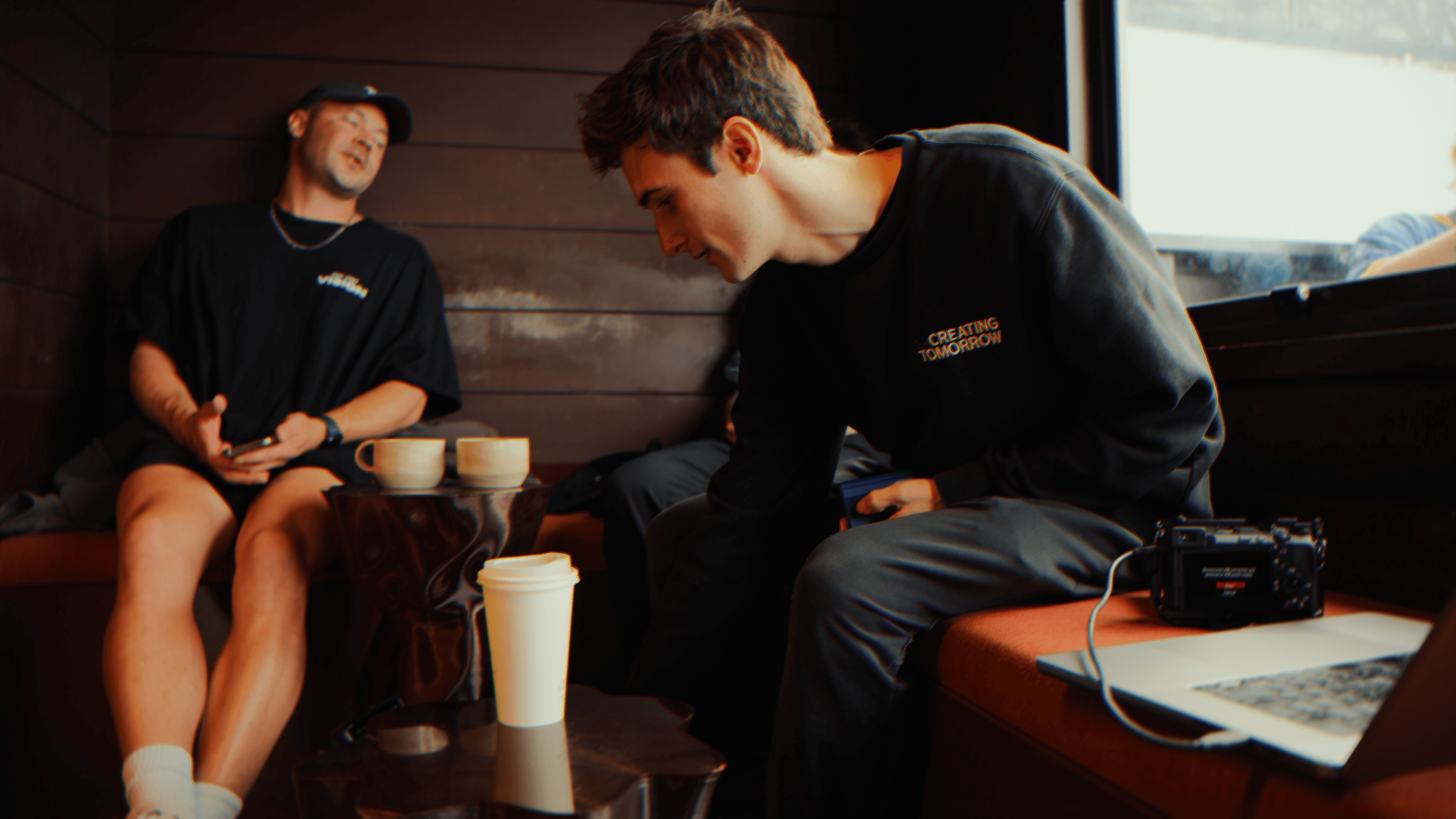 Three people in conversation with warm lighting, including a person wearing a hat and another person wrapped in what appears to be a textured shawl or sweater.