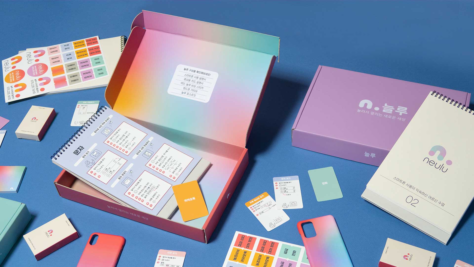 The image shows an open box with a gradient of pink, blue, and green colors, resting on a blue surface. The box contains various items, including a white sheet of paper with Korean text, a yellow sticky note, a small green card, a white notebook with a pink logo, and several colorful stickers scattered around it. To the right of the box is a purple box with the same pink logo and a white notebook resting on top of it. Several small square stickers with Korean text are visible near the bottom right corner of the image. A red phone case is also visible in the bottom left corner.