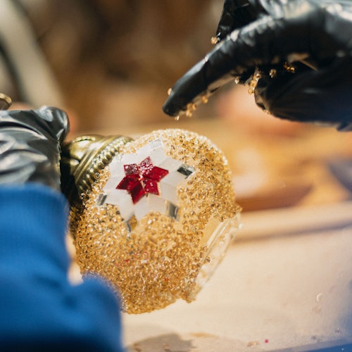 Malapitan ang kamay na nakasuot ng itim na guwantes na nagdekorasyon ng lampara na mosaic gamit ang ginto at pulang mga bato.