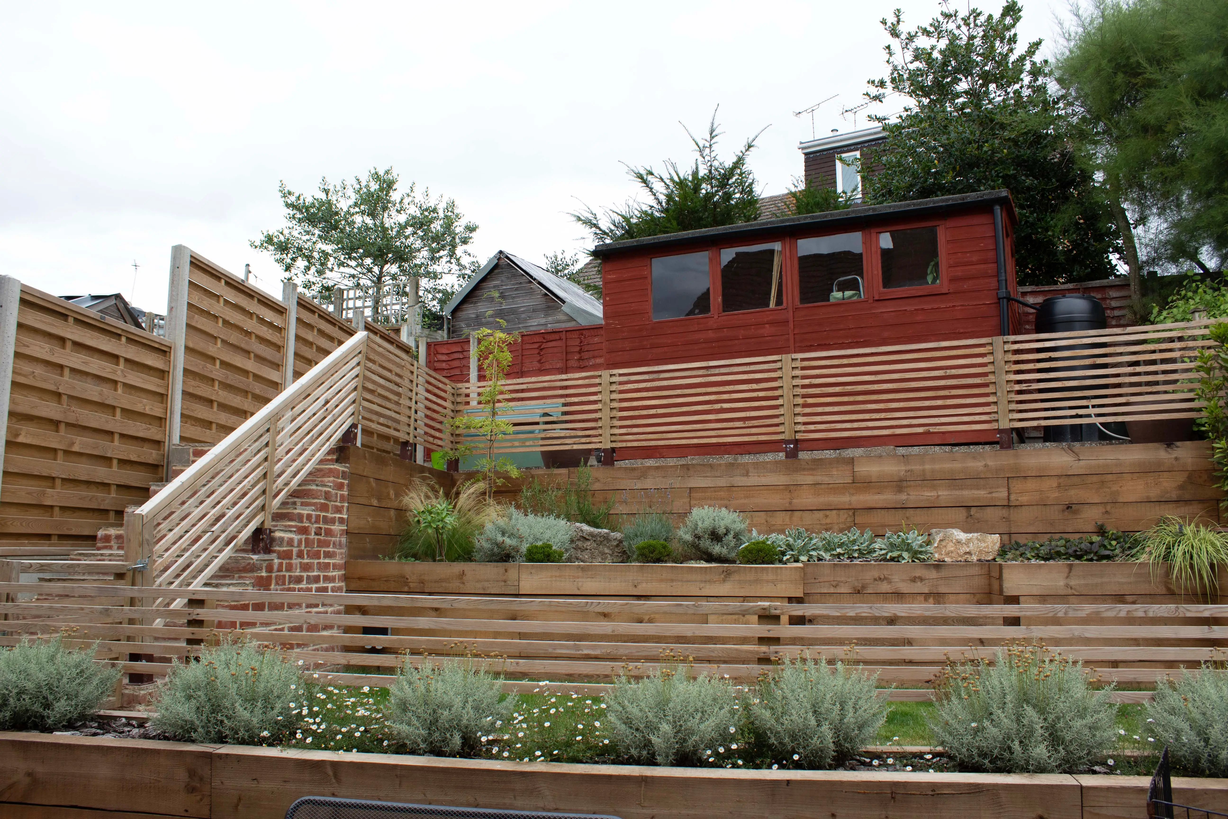 A hillside garden with terraced steps, featuring greenery and a wooden structure at the top against a cloudy sky.