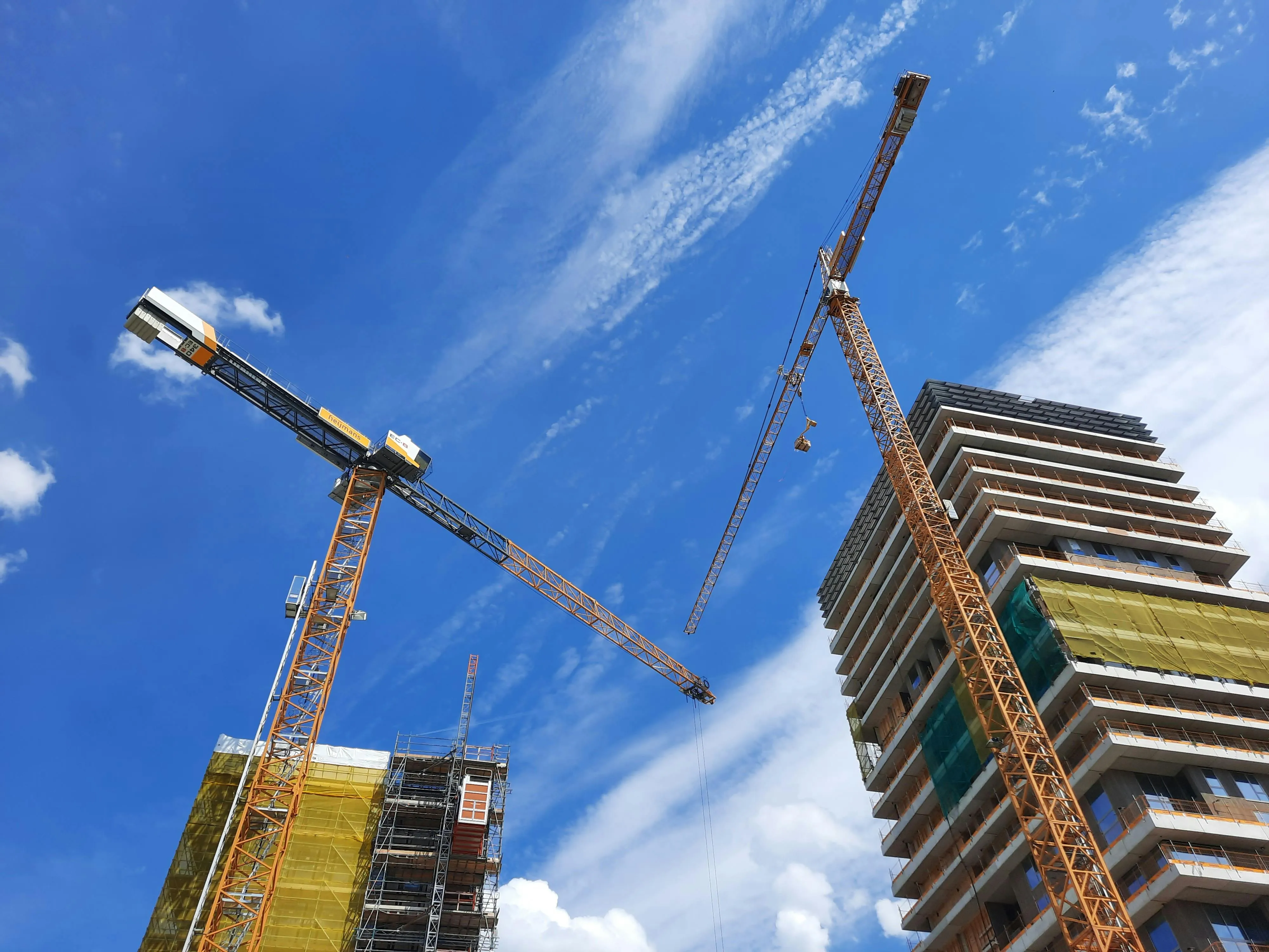Two yellow tower cranes at a high-rise building construction site under a bright blue sky with wispy clouds.