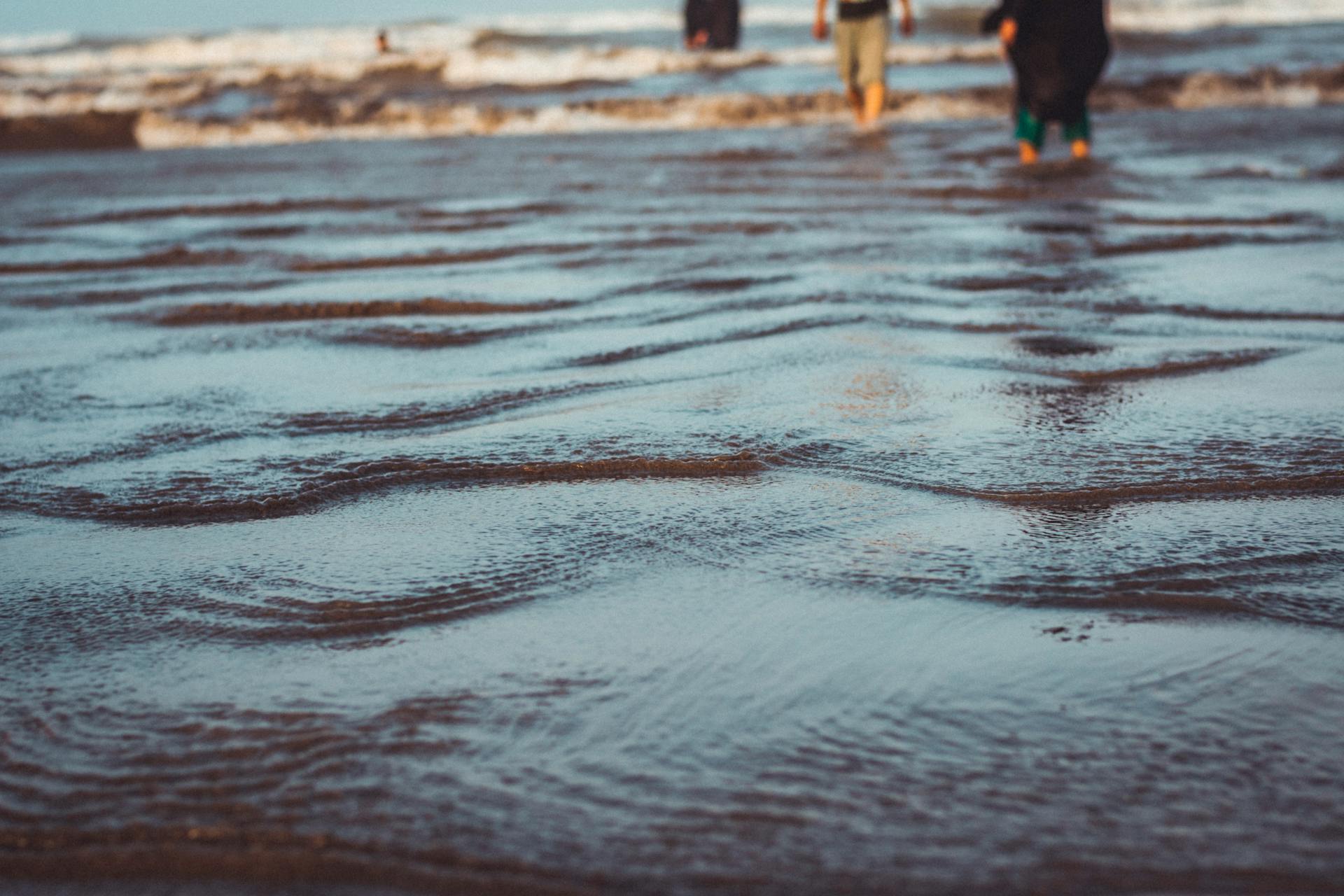 Rippling sand beach with shallow water reflecting the sky. Two people walk toward distant waves, creating a serene and peaceful atmosphere.